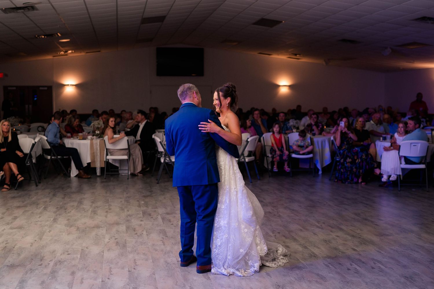 Bride and groom share first dance on dance floor surrounded by seated wedding reception guests.