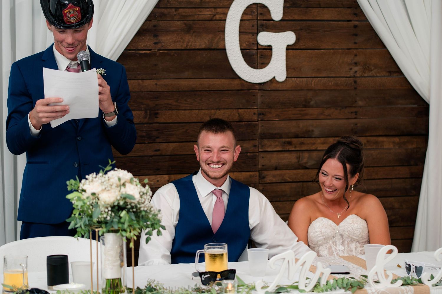 Groom and bride smile as a groomsman in navy suit delivers a speech at the wedding reception.