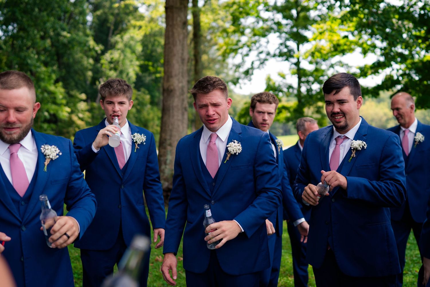 Groomsmen in navy suits and pink ties celebrate outdoors holding champagne bottles before the wedding.