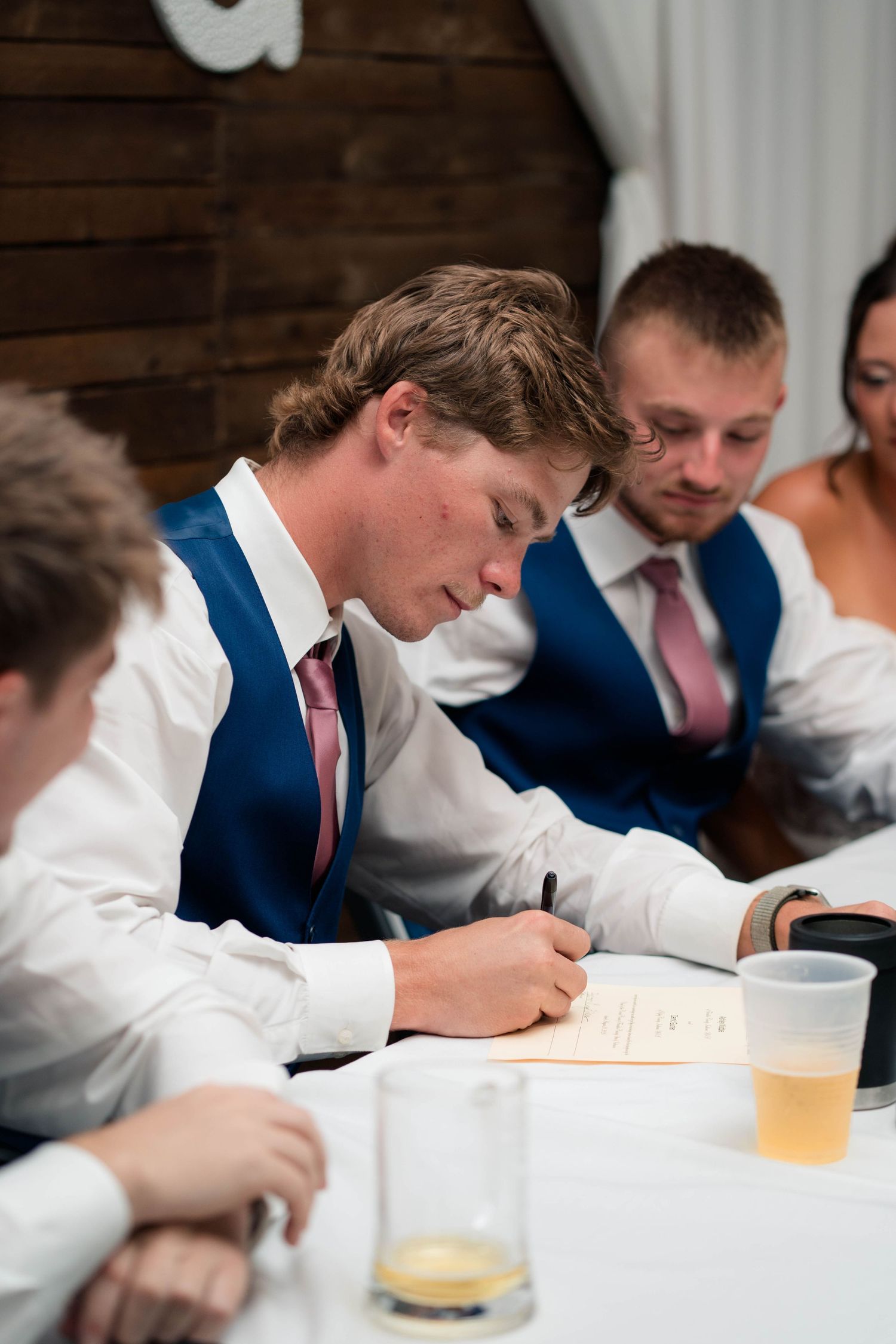 Young man in blue vest and pink tie signing documents at a wedding reception table with guests nearby.