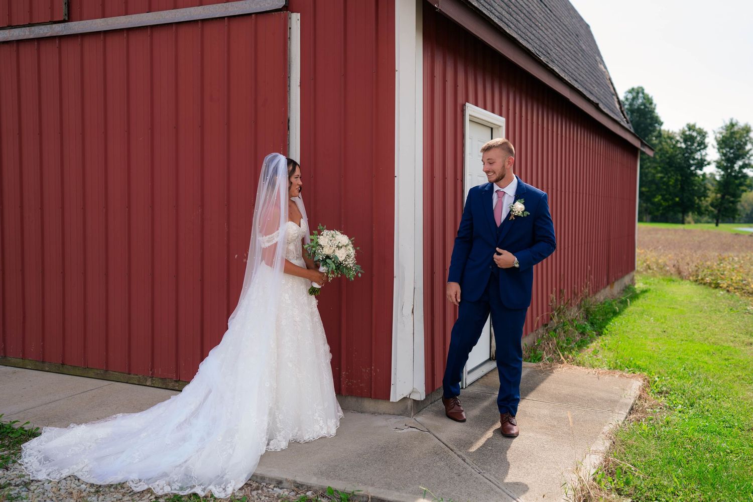 Bride and groom share first look moment outside a red barn, groom in navy suit smiling.