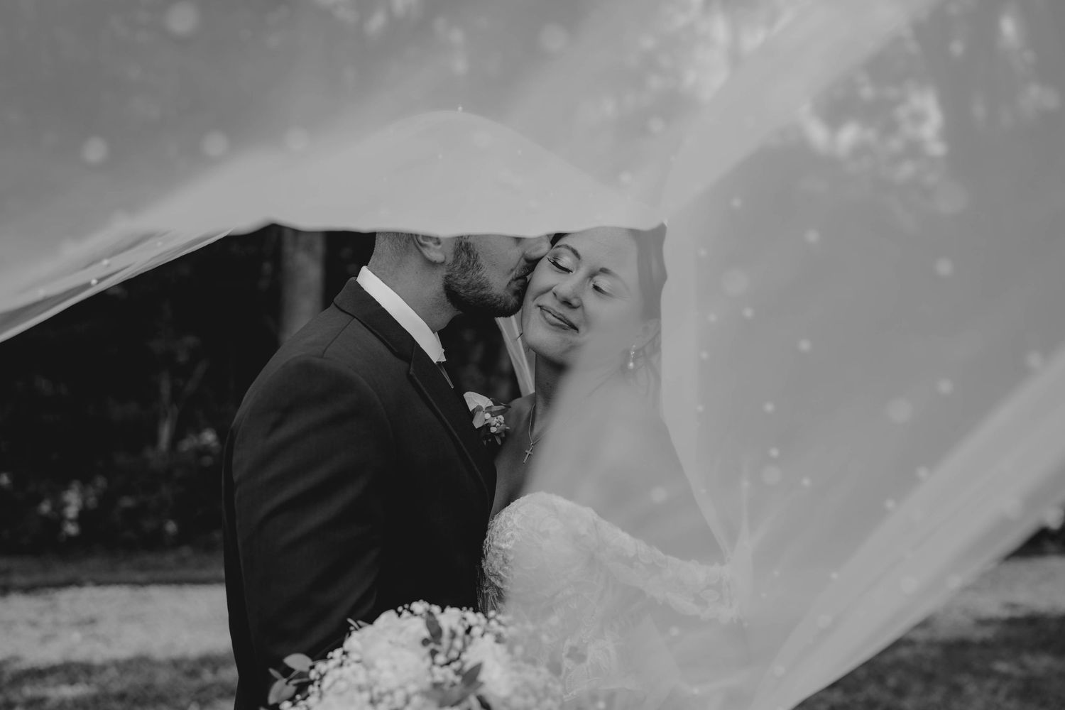 Dramatic black and white wedding photo of groom kissing bride as her veil billows around them in the wind.