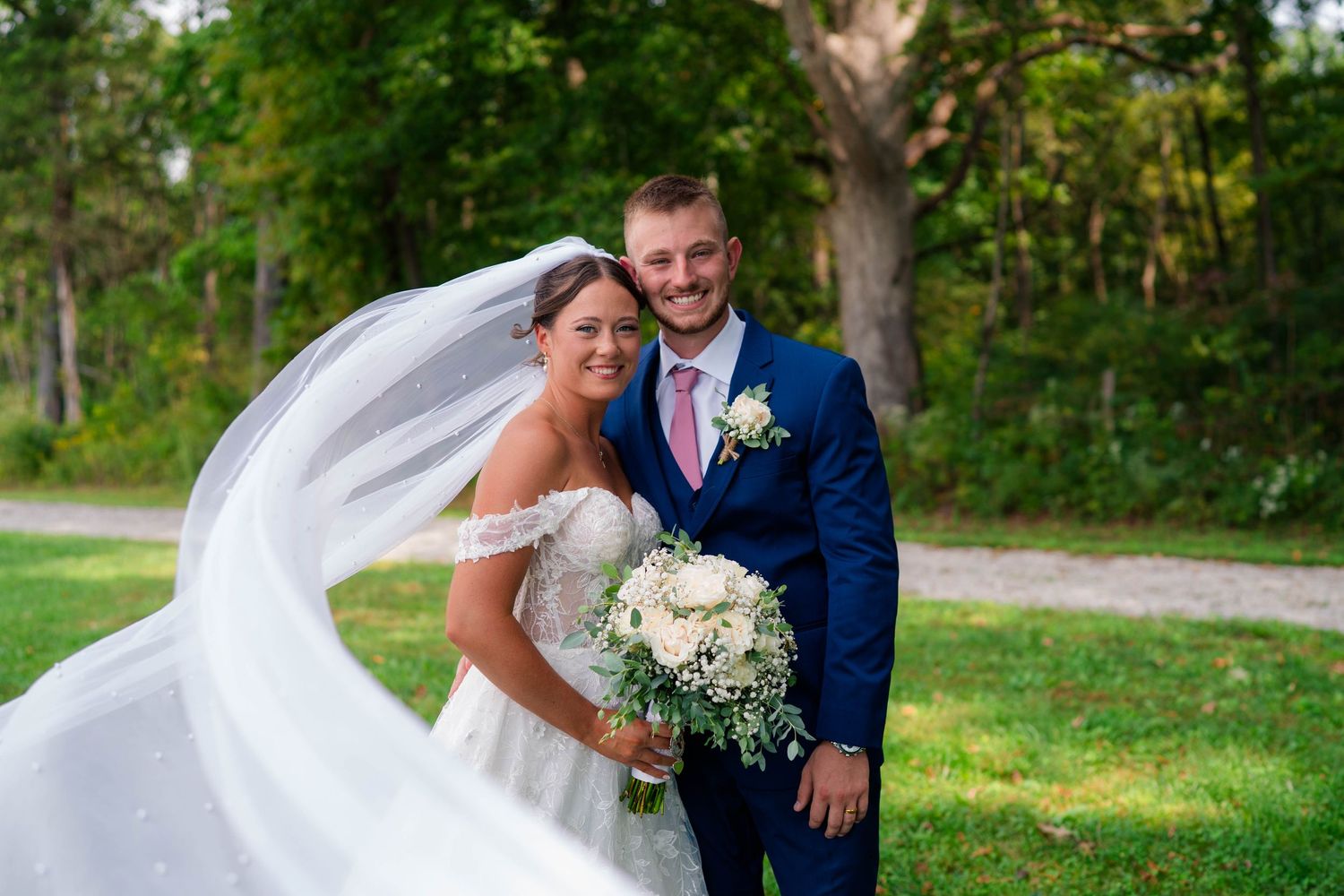 Bride's veil flows in the wind as she and her groom pose together outdoors surrounded by green trees.