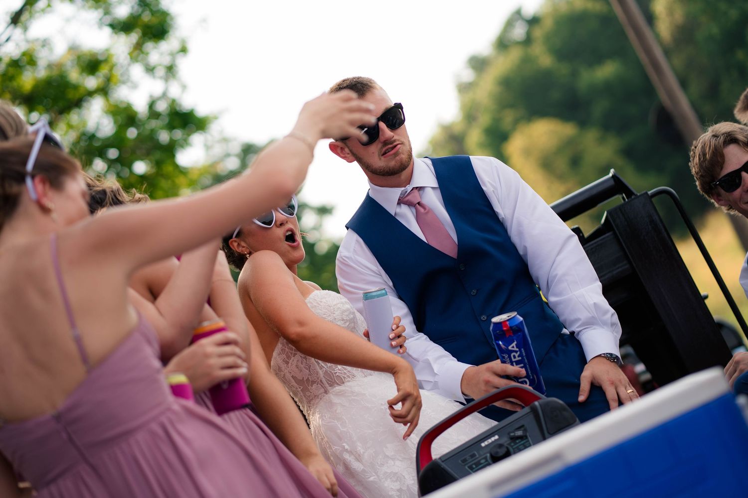 Groom in navy vest and pink tie reaches for a drink at lively wedding reception.