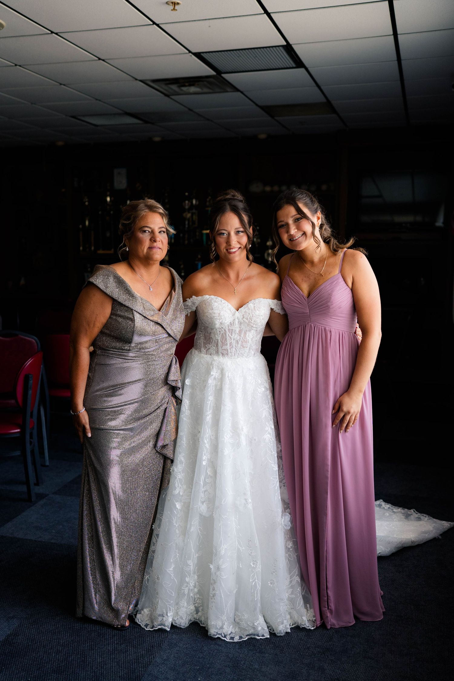 Bride in white lace off-shoulder gown posing with two women in mauve bridesmaid dresses.