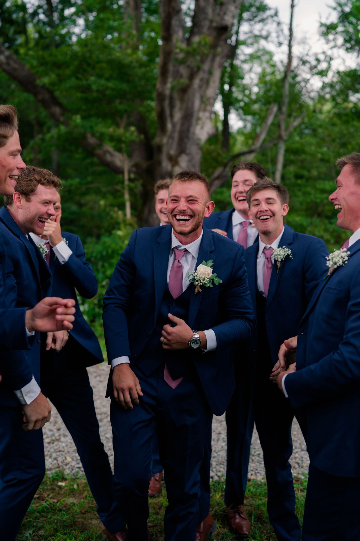 Groomsmen in navy suits with pink ties laughing together outdoors at a wedding celebration.