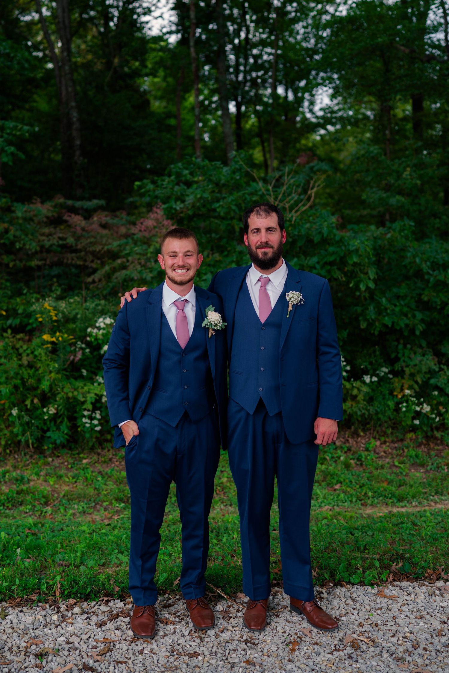 Two groomsmen in navy blue suits with pink ties pose outdoors at a wedding.