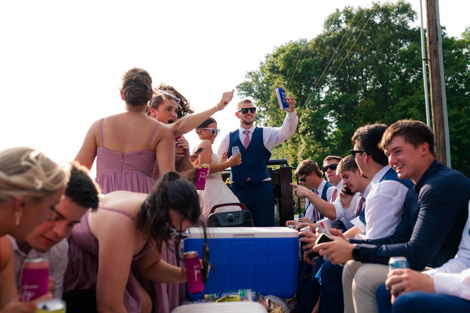 Groom raises bottle while wedding party celebrates on truck bed with bridesmaids in mauve dresses outdoors.