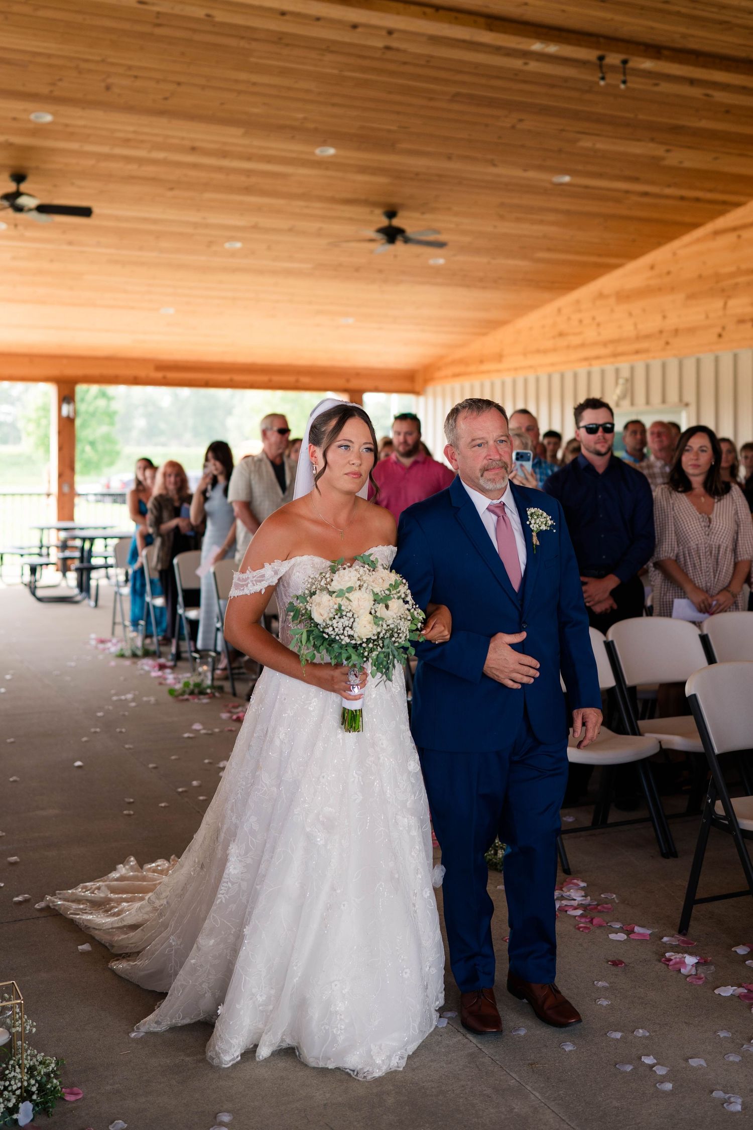 Bride in off-shoulder lace gown walks down the aisle with escort during outdoor wedding ceremony.