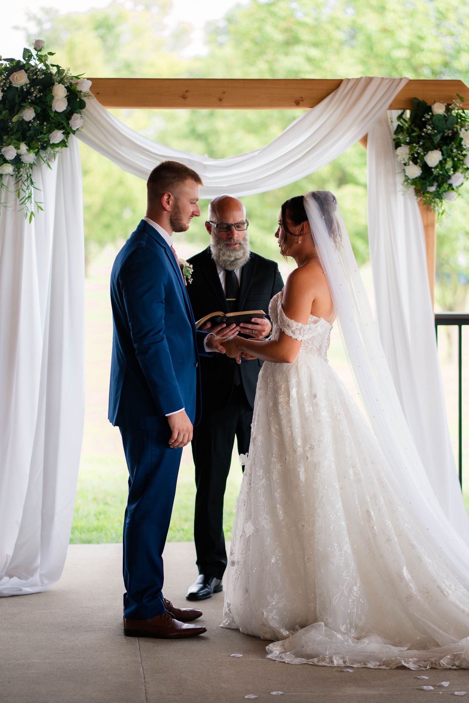 Groom and bride exchange rings with officiant under a decorated wooden arch at outdoor ceremony.