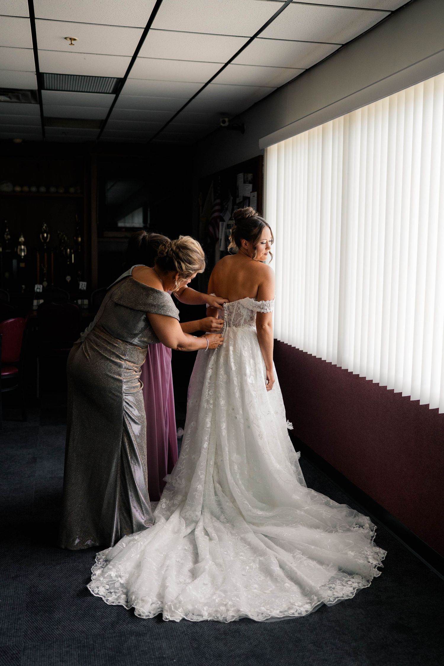A bride in a lace off-shoulder gown gets her dress fastened by a woman in silver near a bright window.