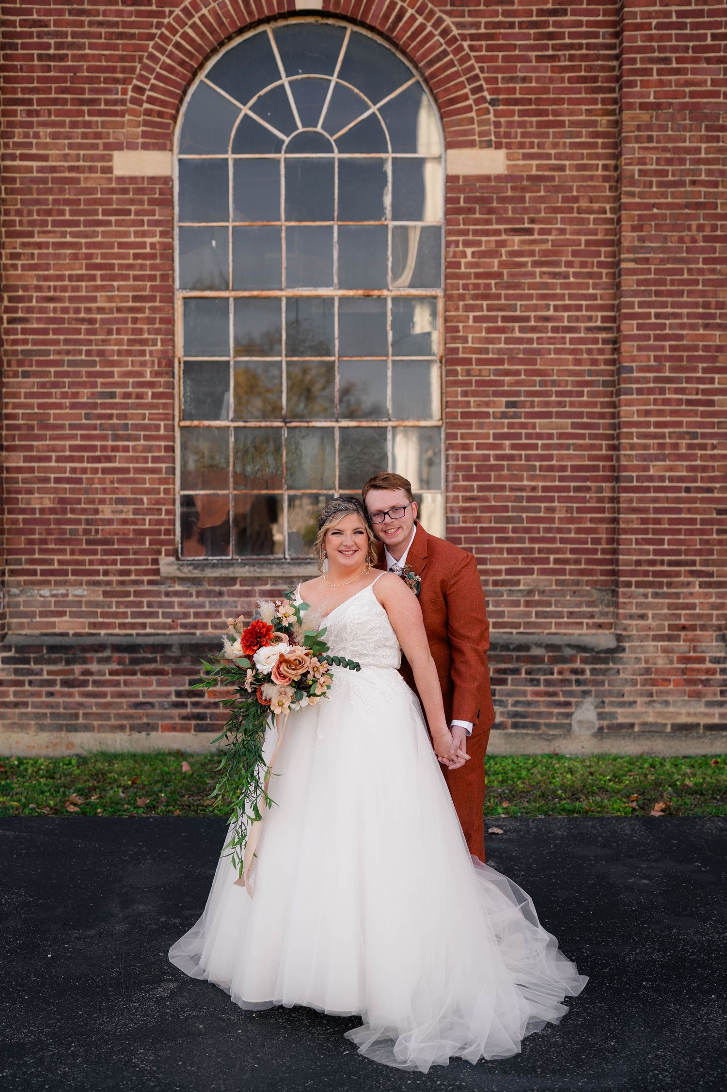 Bride and groom smiling together in front of rustic brick building with large arched windows.
