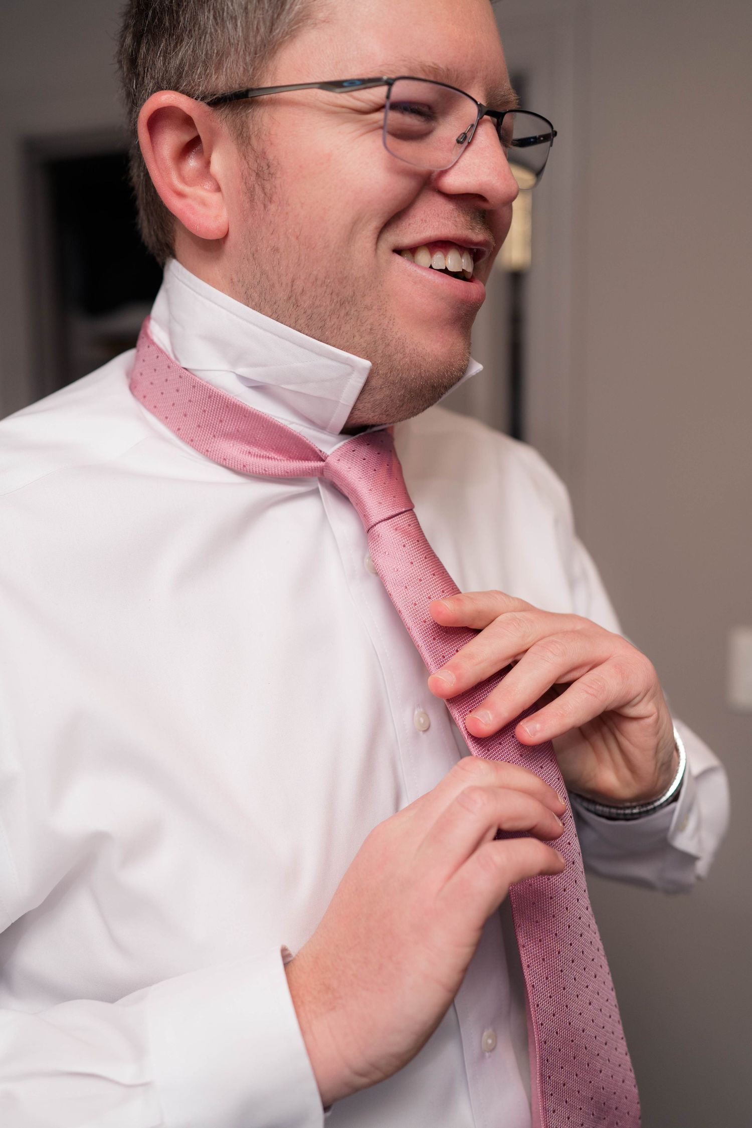 Smiling man with glasses adjusting a pink tie over a white dress shirt.