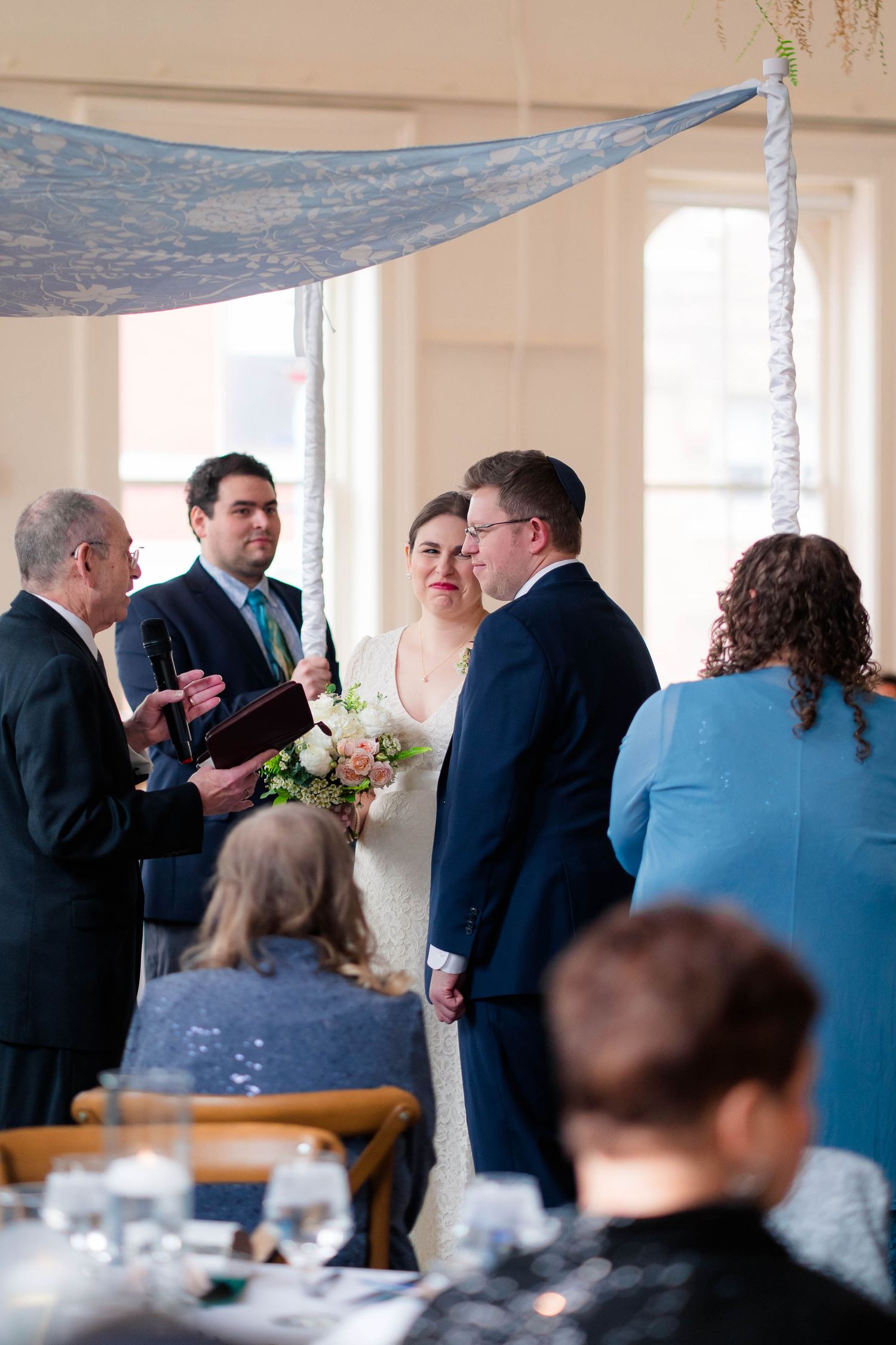 Bride and groom exchange vows under blue chuppah with officiant and wedding party present.