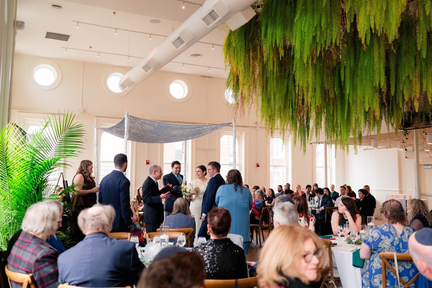 Jewish wedding ceremony under chuppah with hanging ferns and guests seated in bright venue.