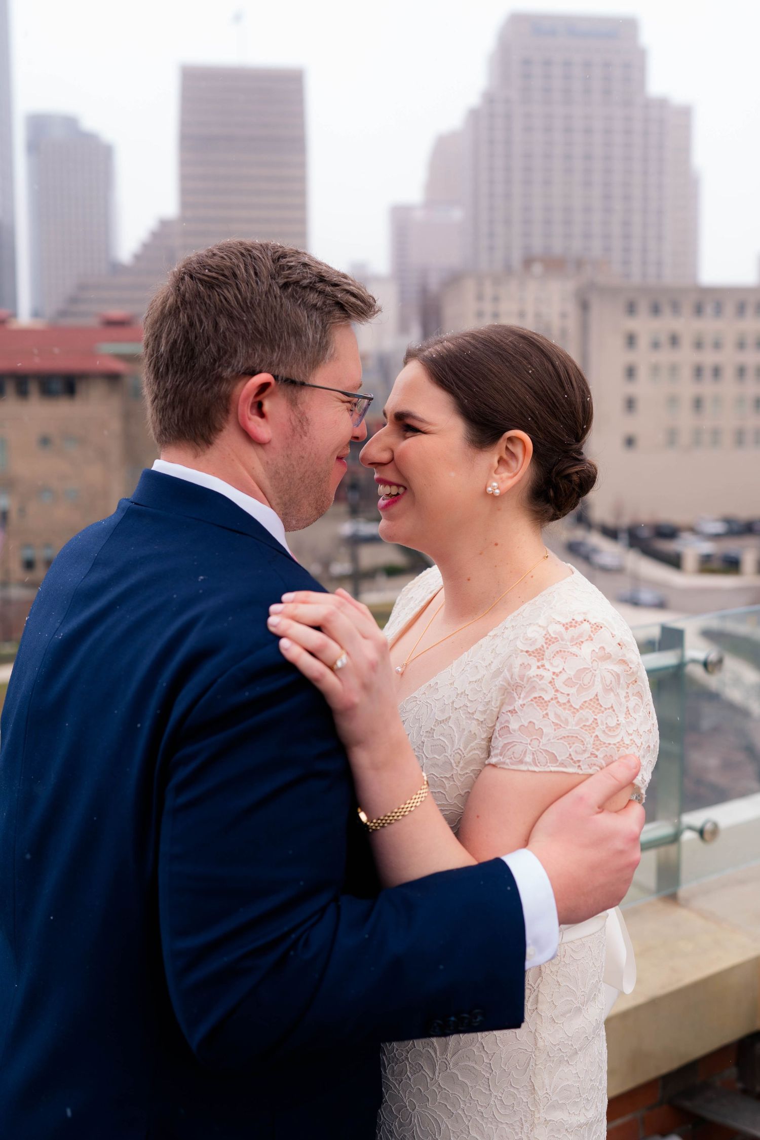 Laughing bride and groom share a joyful embrace on a rooftop with a foggy urban skyline behind them.