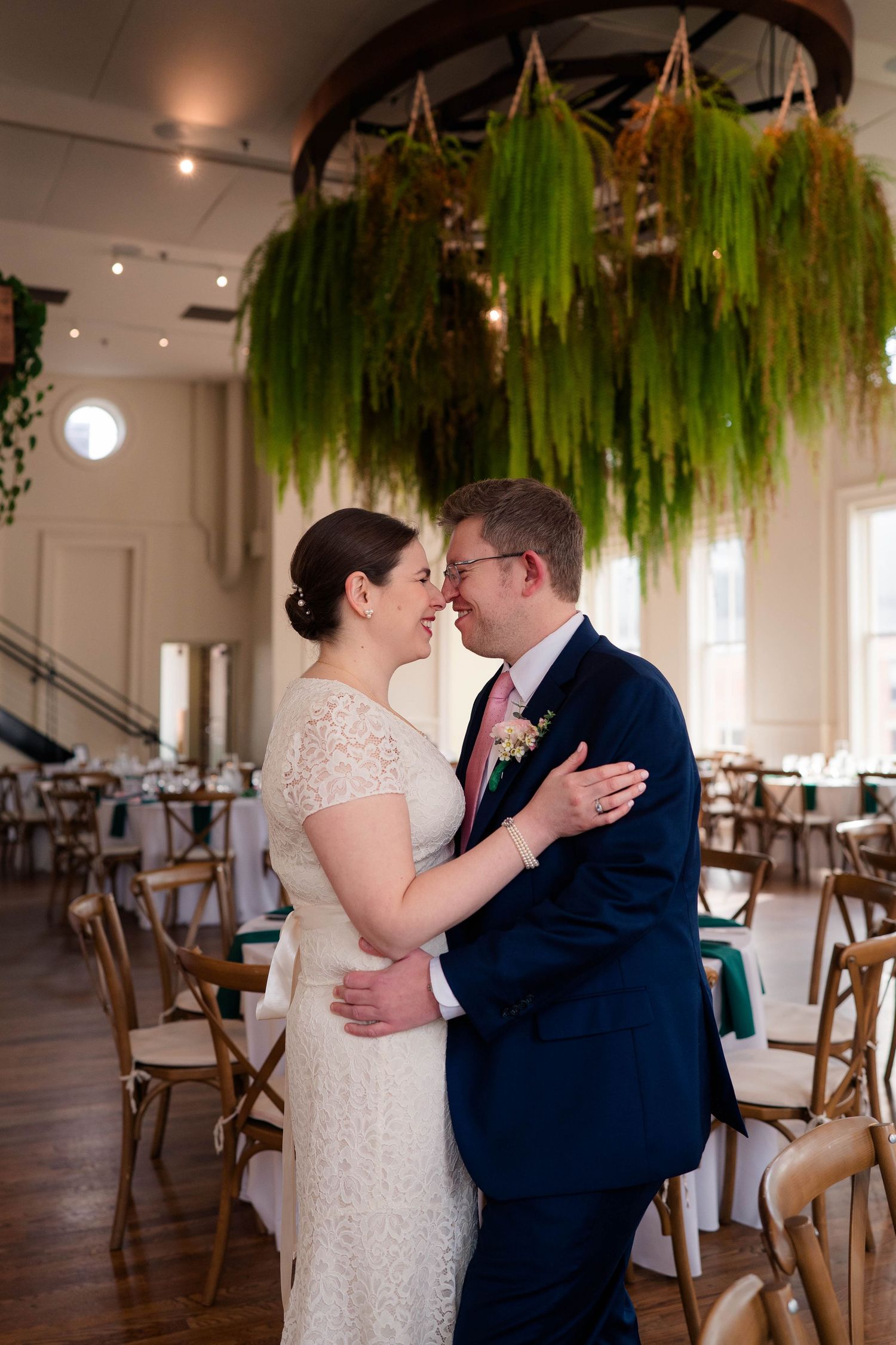 Bride and groom sharing an intimate nose-to-nose moment beneath a lush hanging greenery display at their wedding.