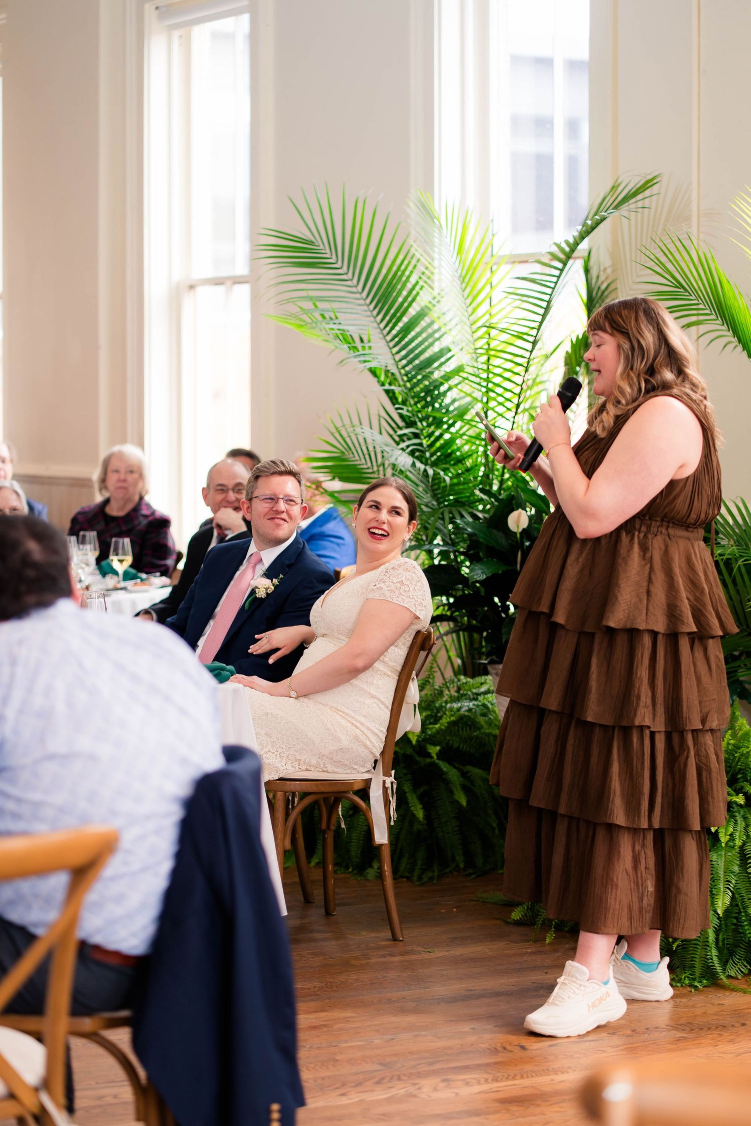 Wedding speaker in brown dress making guests laugh during a heartfelt reception toast speech.