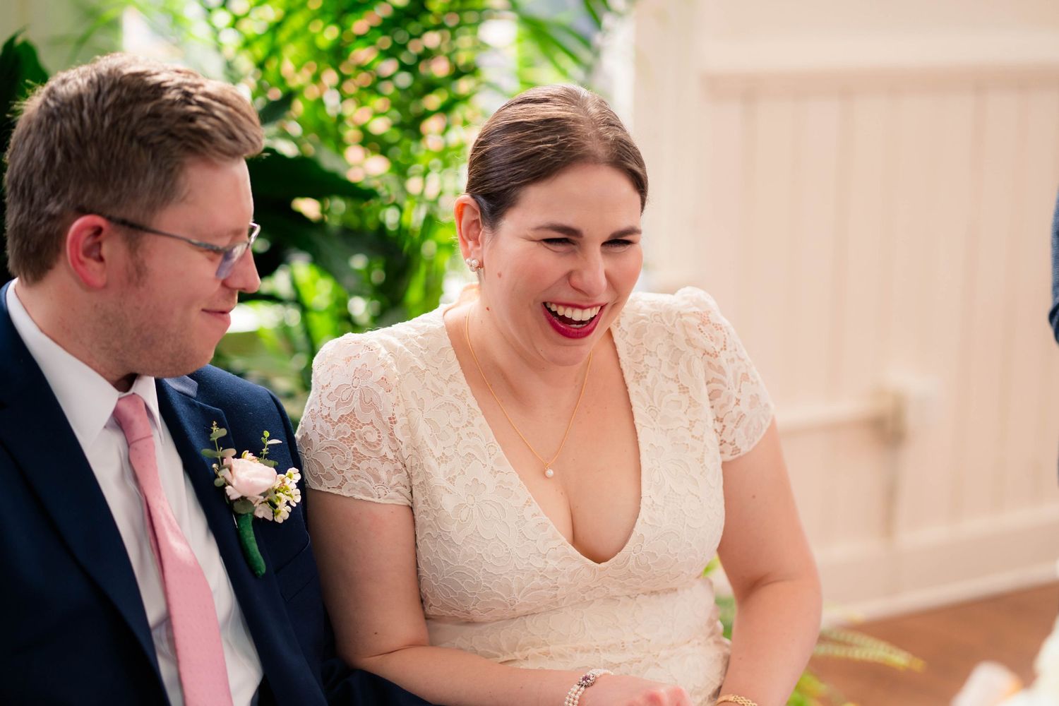 Bride laughs joyfully in white lace dress beside groom in navy suit at reception.