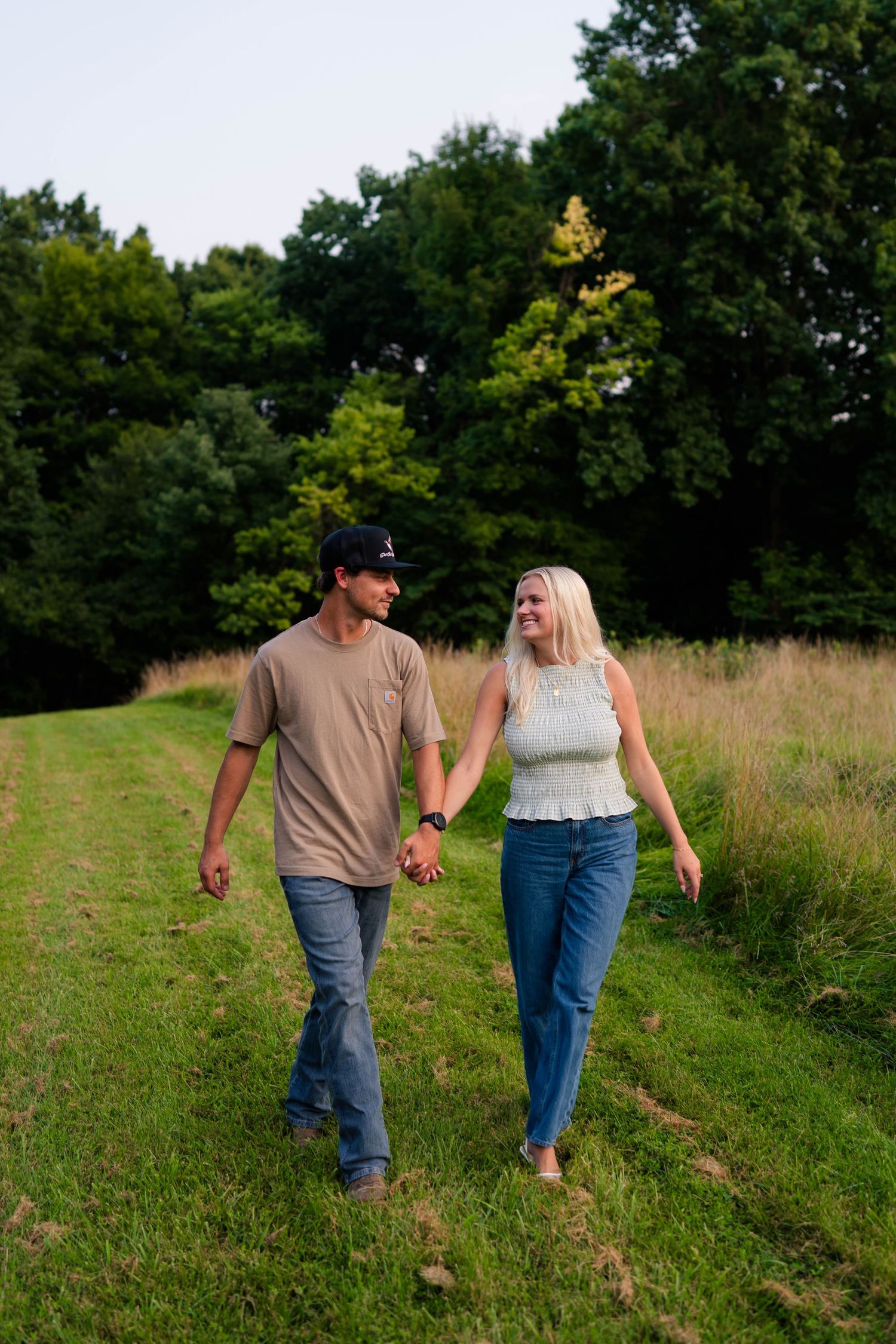 A couple walks hand in hand along a grassy path surrounded by lush green trees in warm evening light.