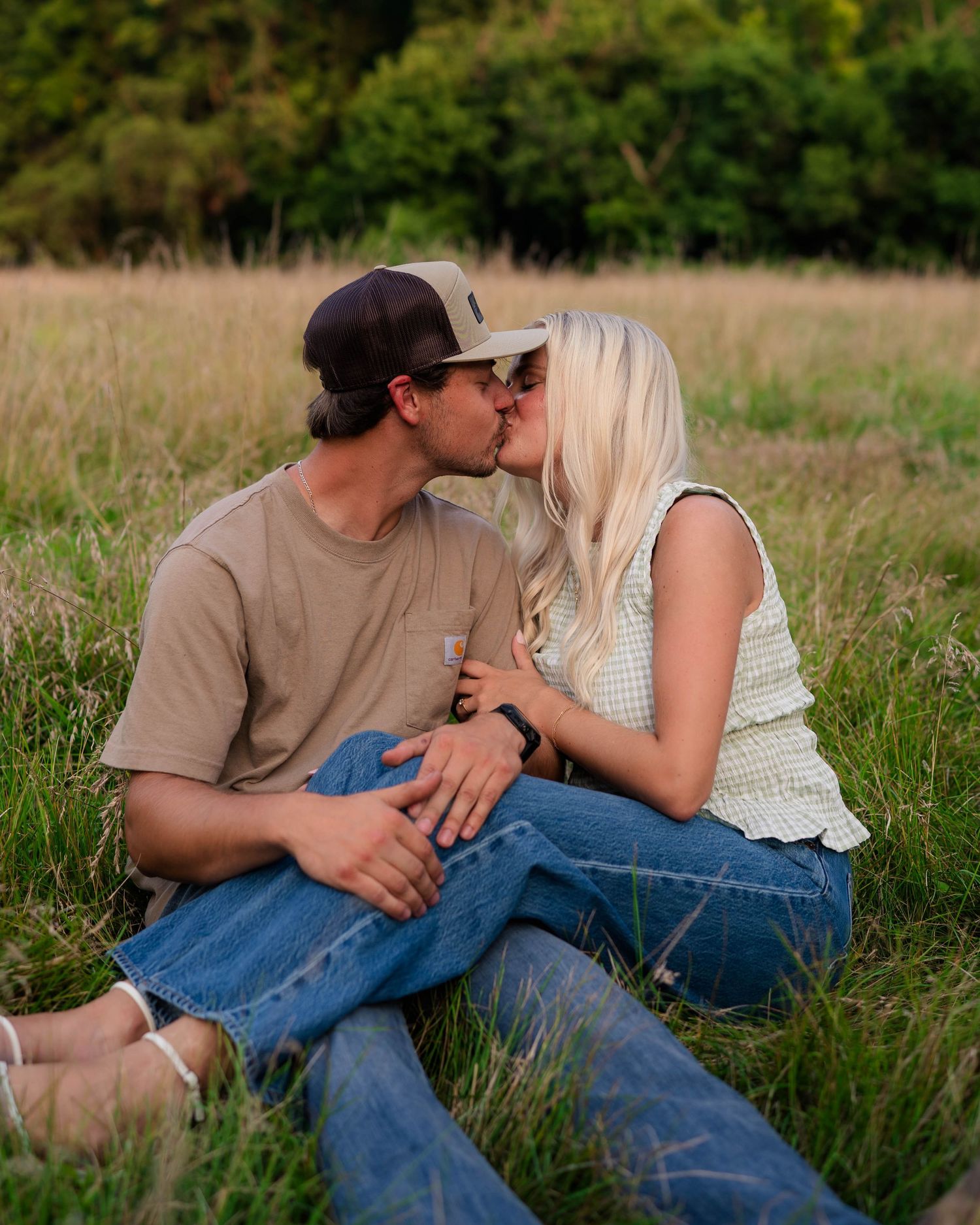 A couple sits in a grassy field, sharing a kiss, with the woman in a white top and man in a tan shirt and cap.