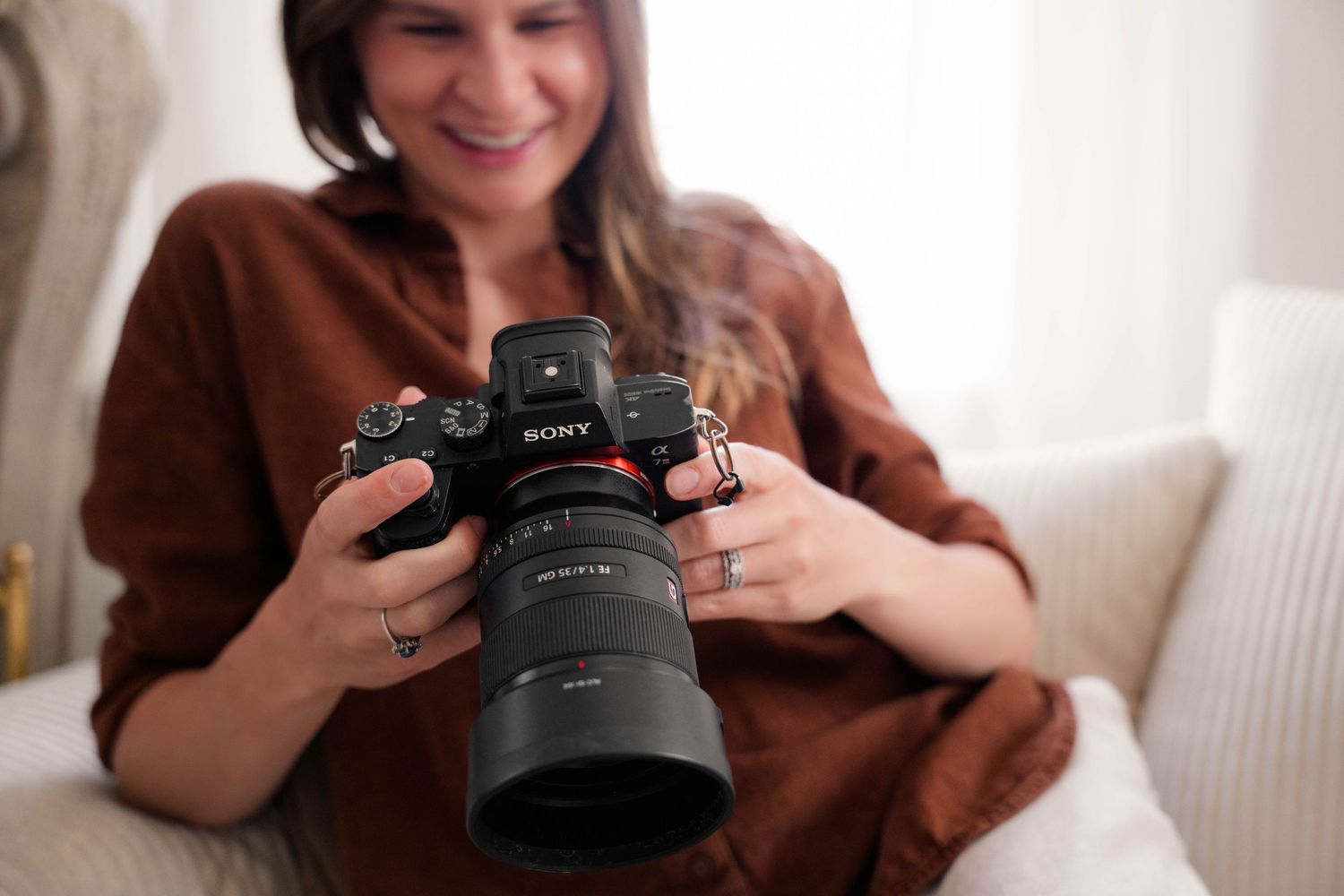 Smiling woman holding Sony mirrorless camera indoors in brown top.