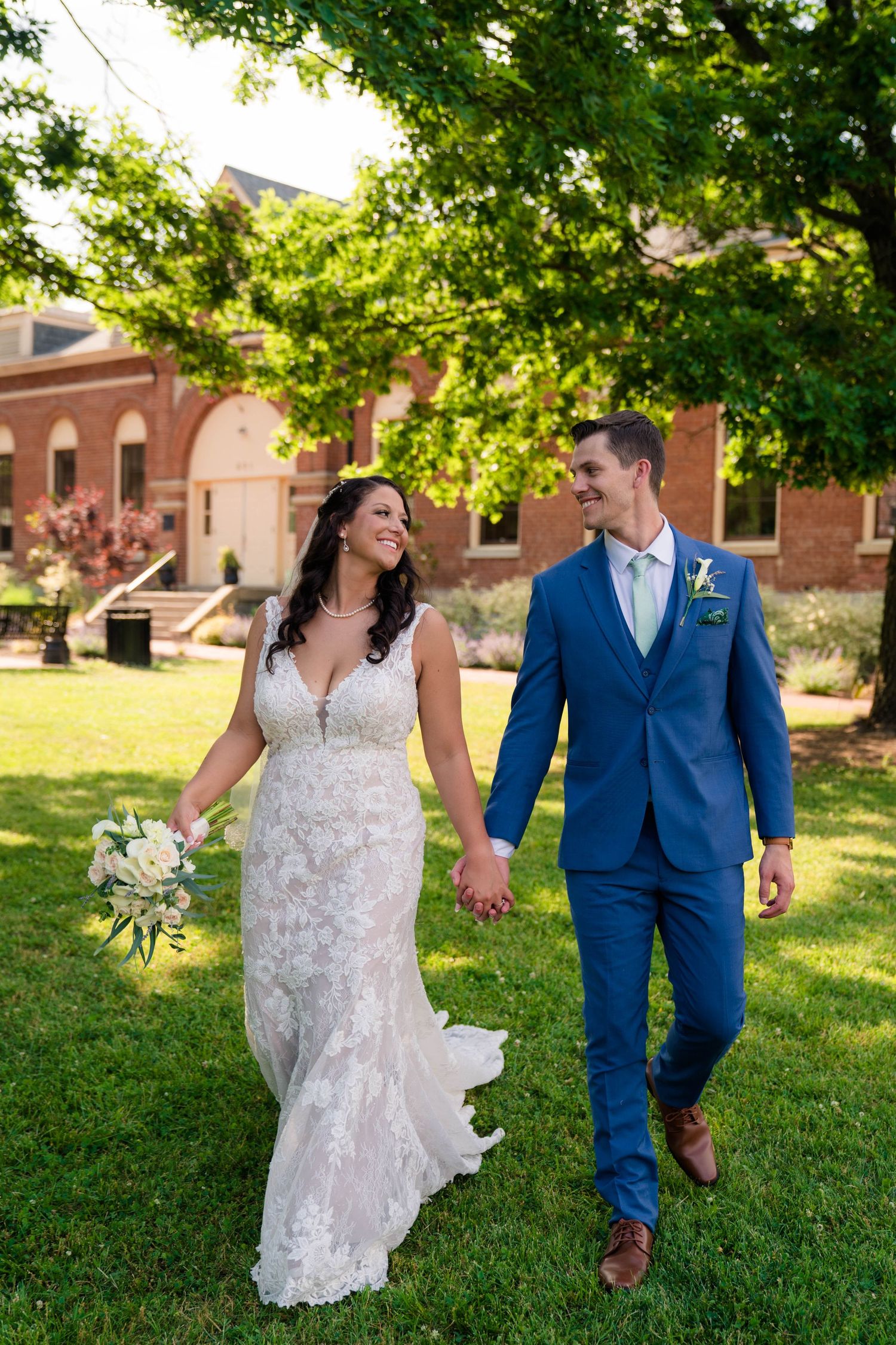 Smiling bride and groom in blue suit stroll hand-in-hand on green lawn outdoors.