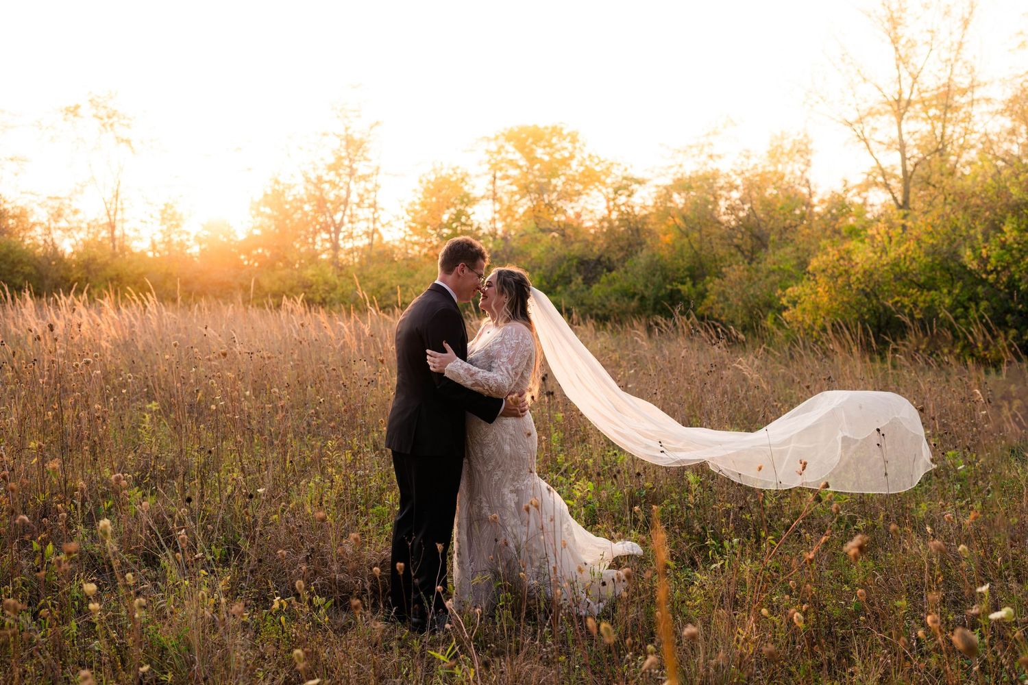 Bride and groom embrace in golden sunset light in meadow with veil flowing in the wind.