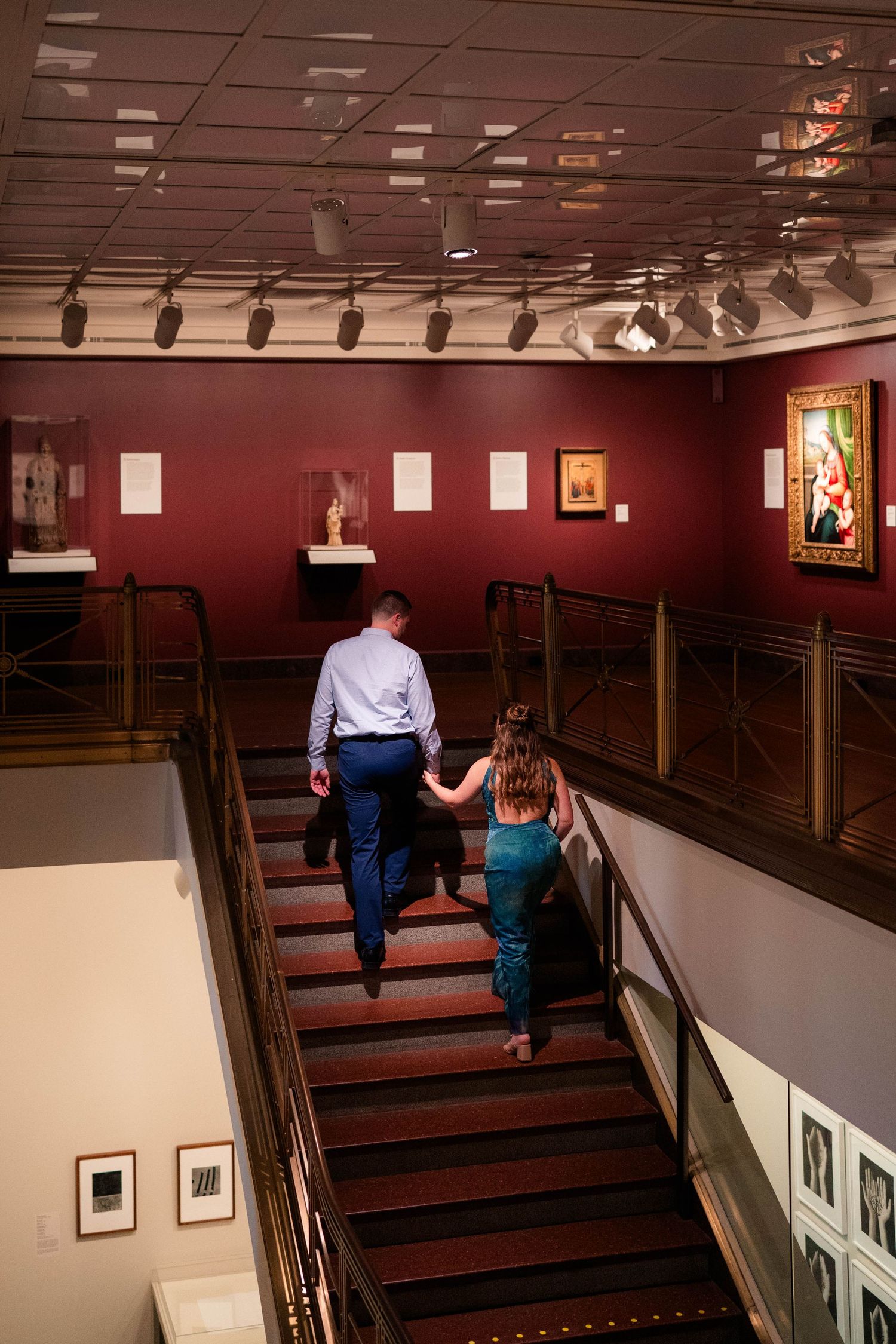 A couple holds hands while climbing stairs inside a museum with red walls and artwork.