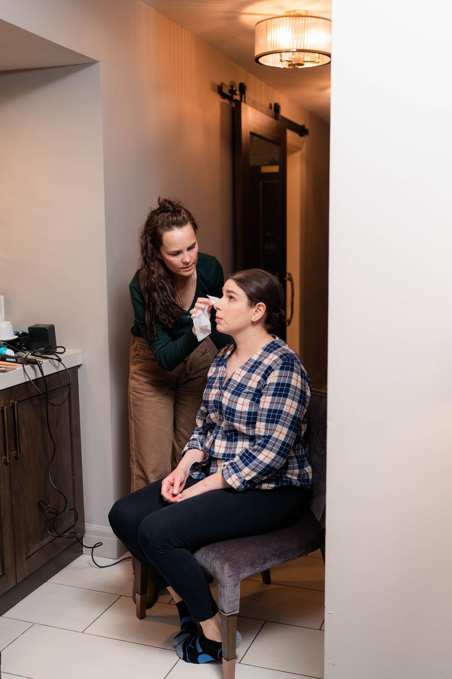 Makeup artist applying cosmetics to a seated woman in a plaid shirt before a wedding.