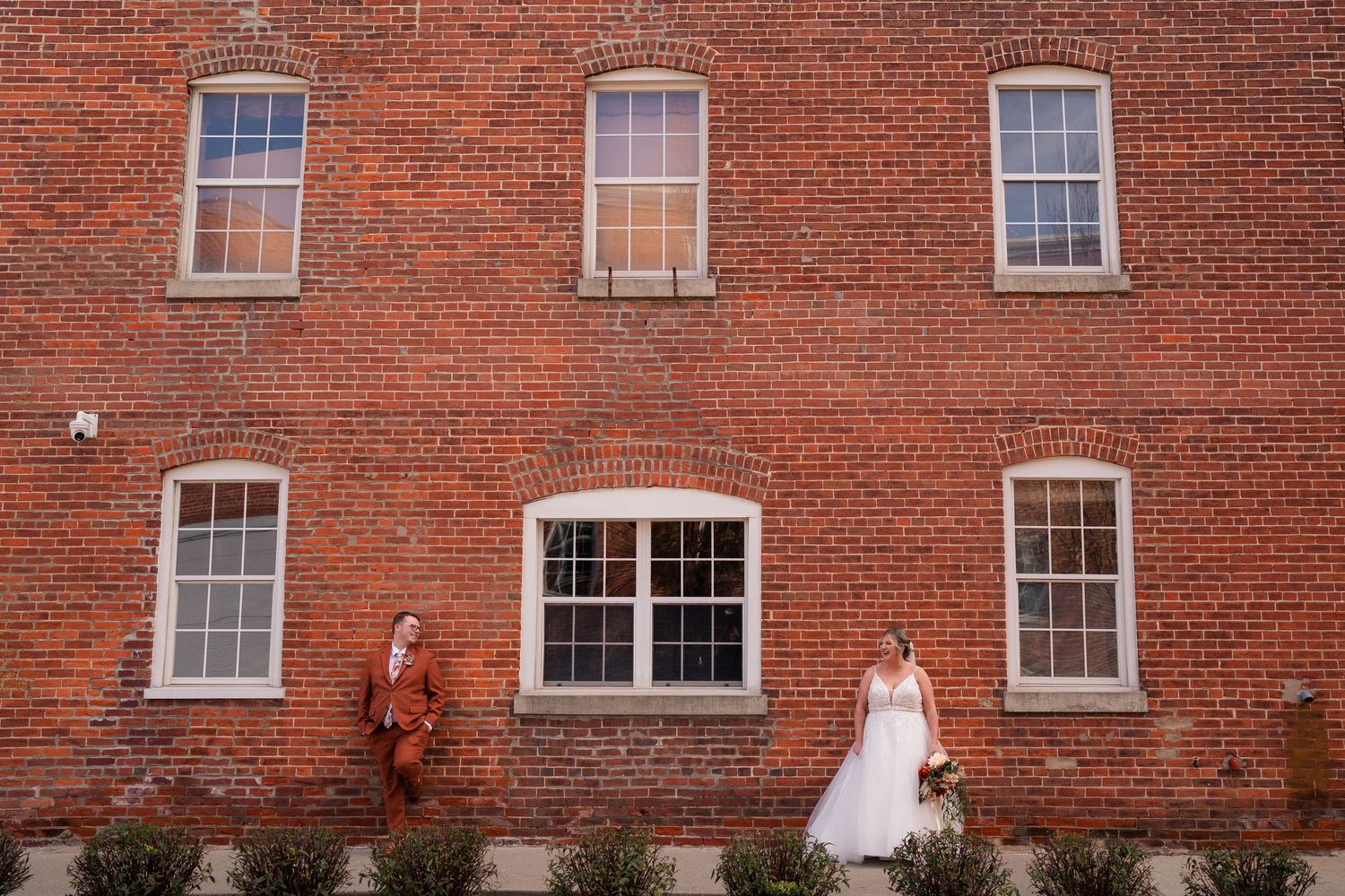 Bride and groom pose apart against a rustic red brick building wall during their wedding day portrait session.