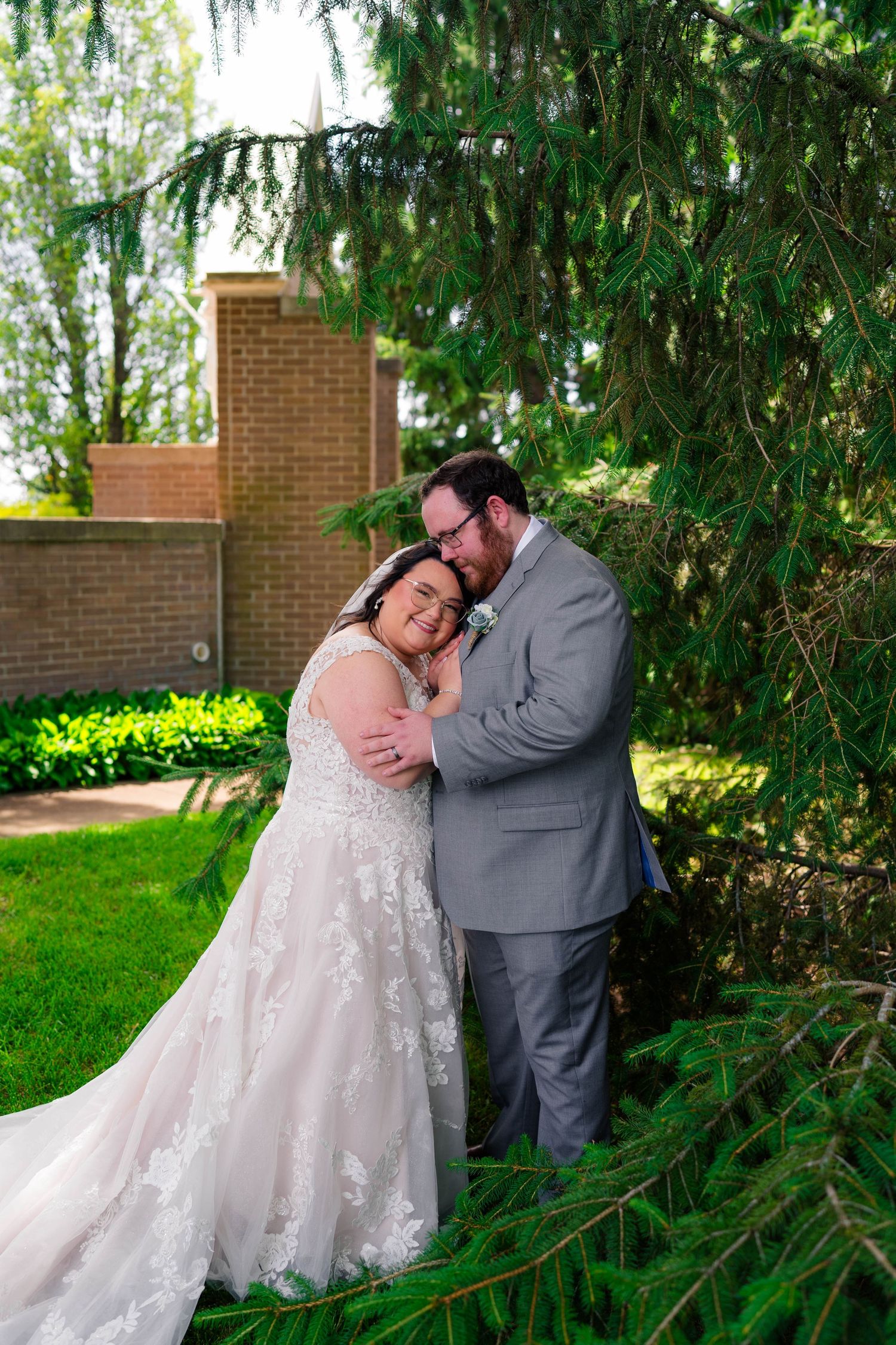 Bride and groom embrace tenderly under pine tree in sunlit garden after wedding ceremony.