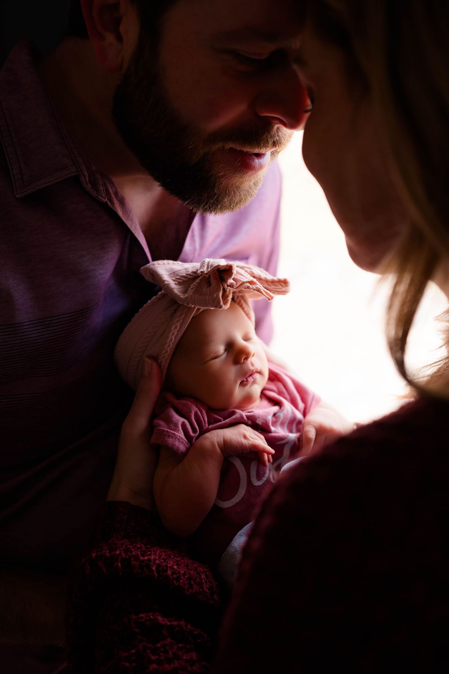 New parents gaze lovingly at their sleeping newborn baby girl wearing a pink bow headband.