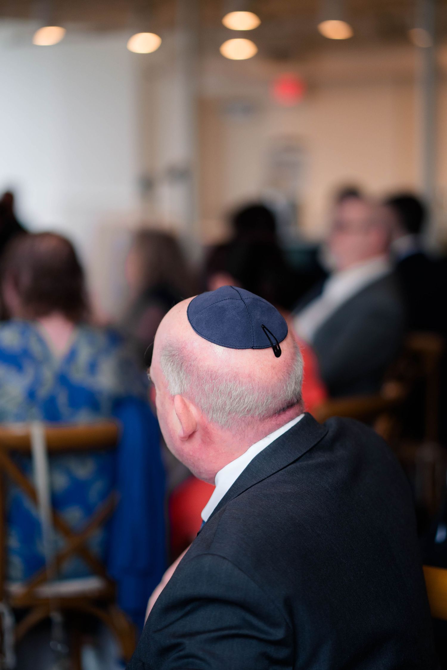 Bald man wearing a navy blue kippah at a Jewish wedding ceremony, seated among other guests.
