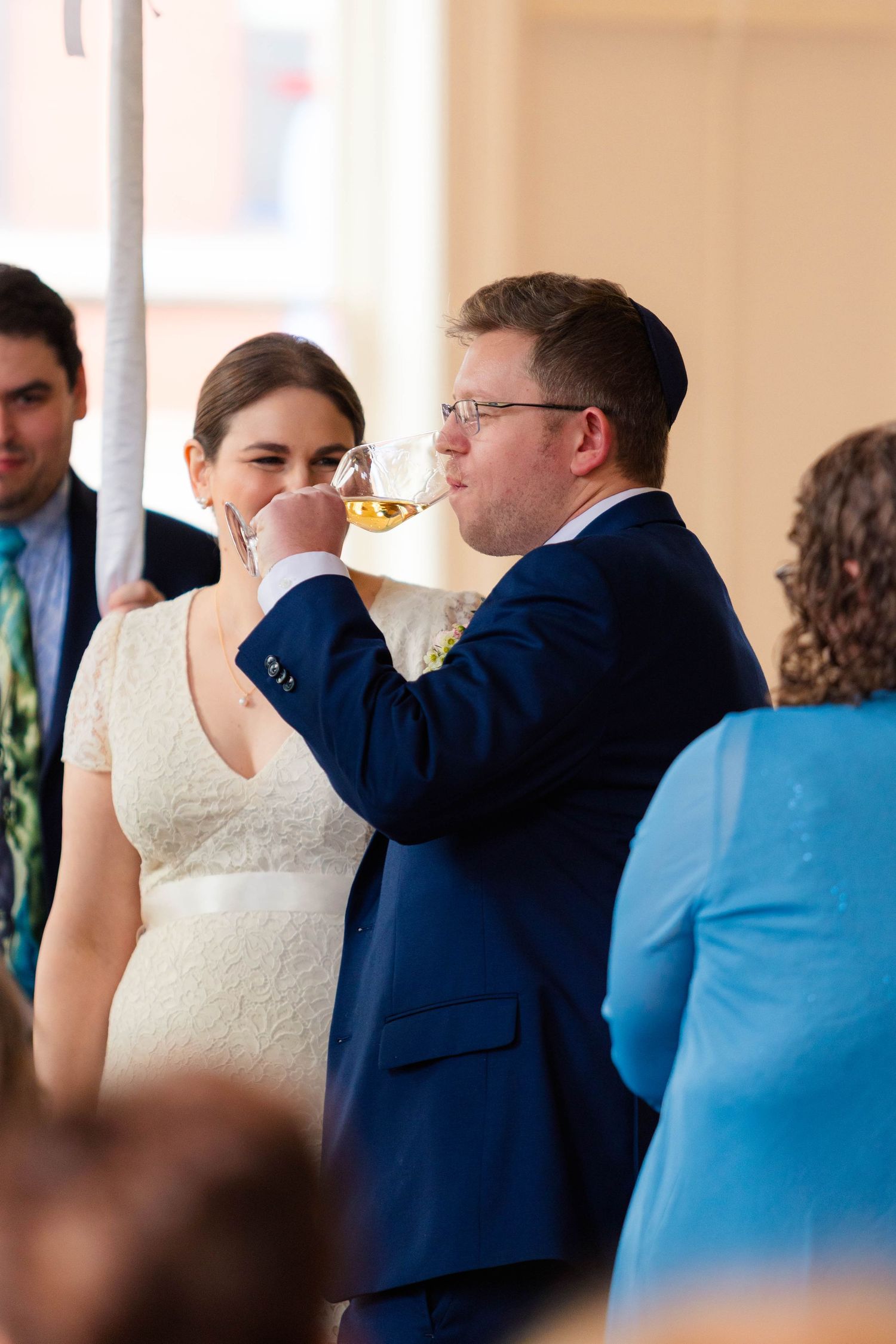 Groom in navy suit drinks from wine glass beside smiling bride in lace dress at wedding.