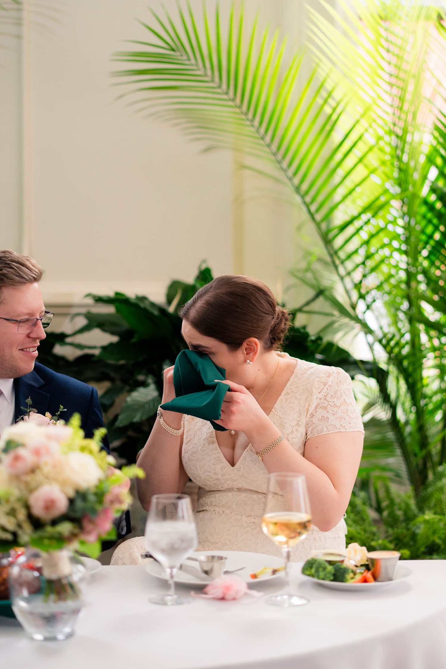 Emotional bride covering her face with a green napkin while laughing at the wedding reception table.