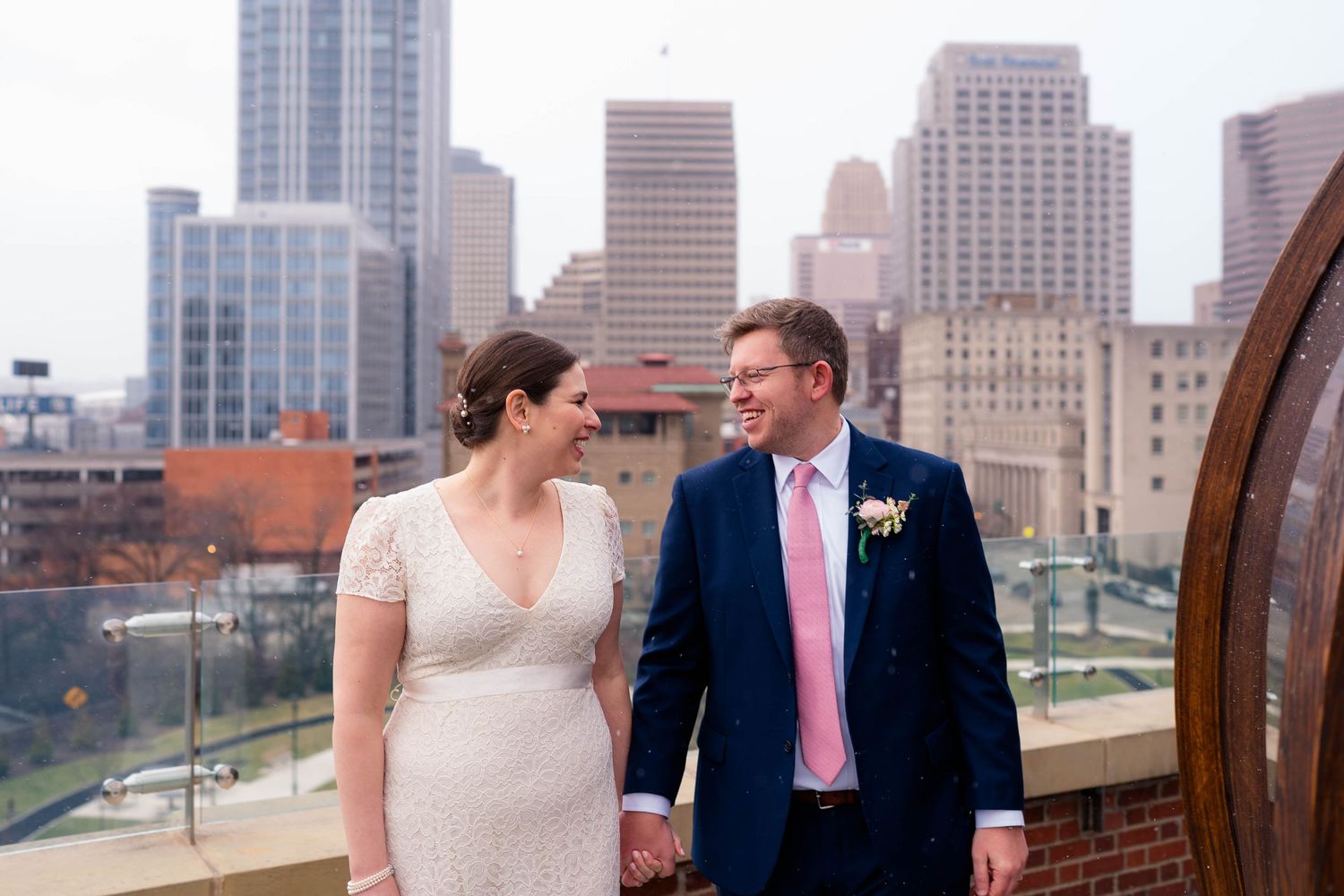 Bride and groom holding hands smiling on rooftop with city skyline backdrop on their wedding day.