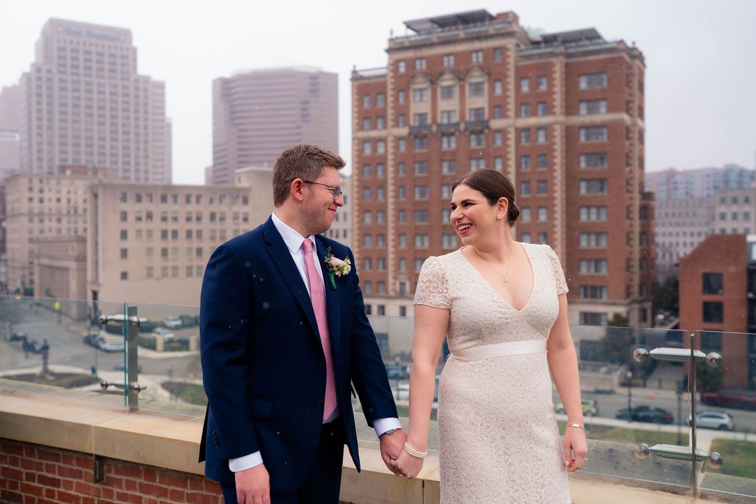 Smiling bride and groom hold hands on a rooftop terrace with a classic brick city building backdrop.