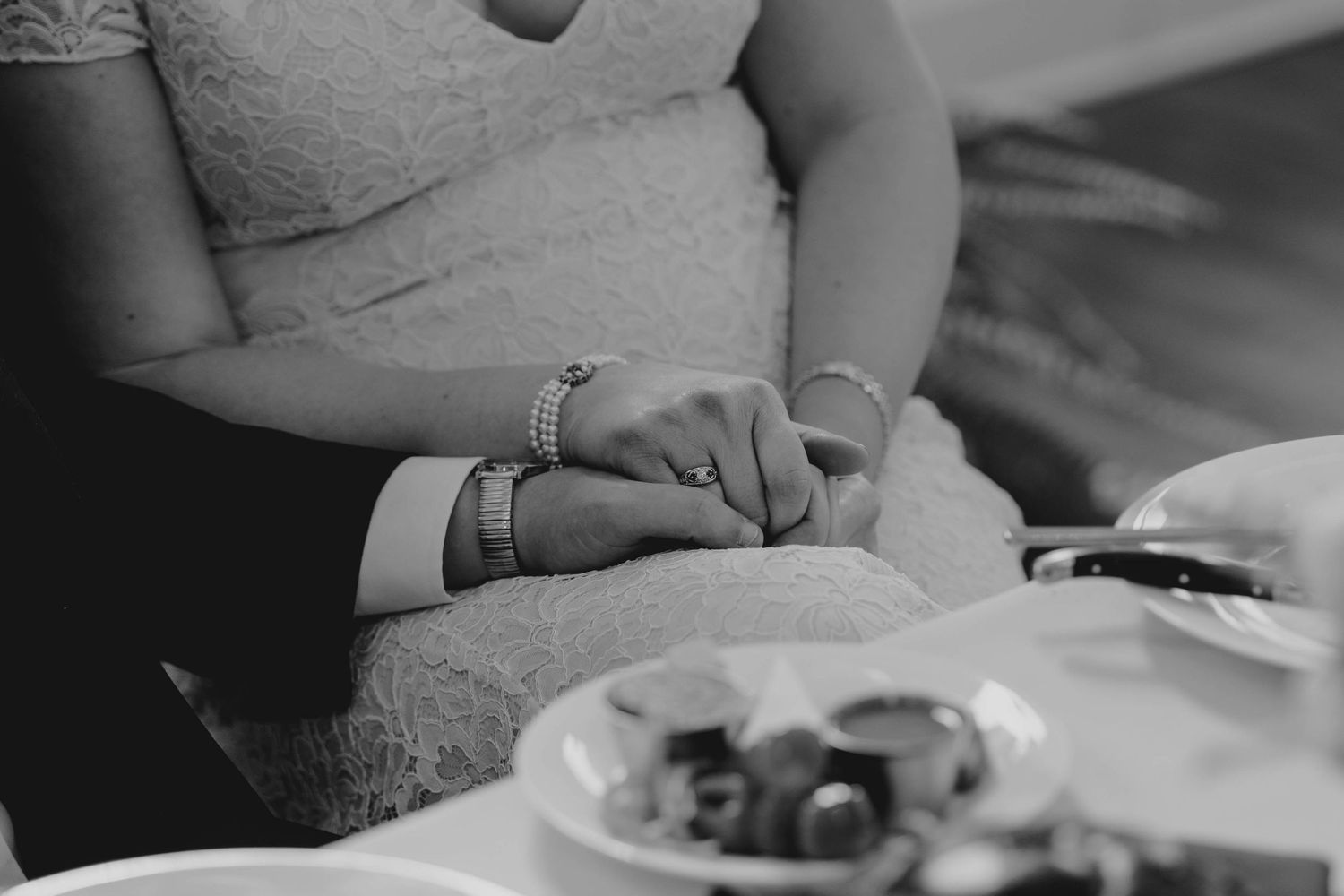 Black and white close-up of bride and groom holding hands at wedding reception table.