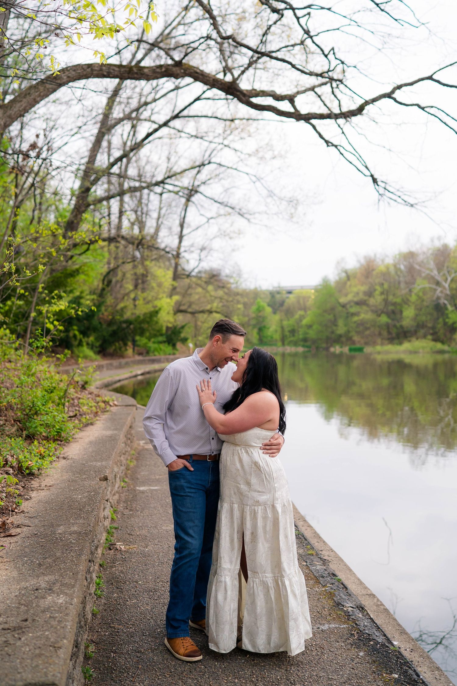 Engaged couple sharing a romantic moment on a lakeside path surrounded by spring foliage.