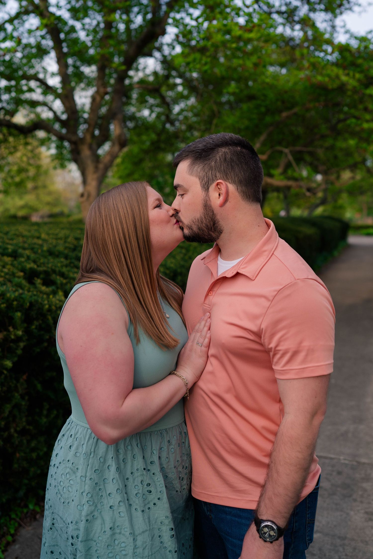 Couple sharing a romantic kiss outdoors surrounded by green hedges.