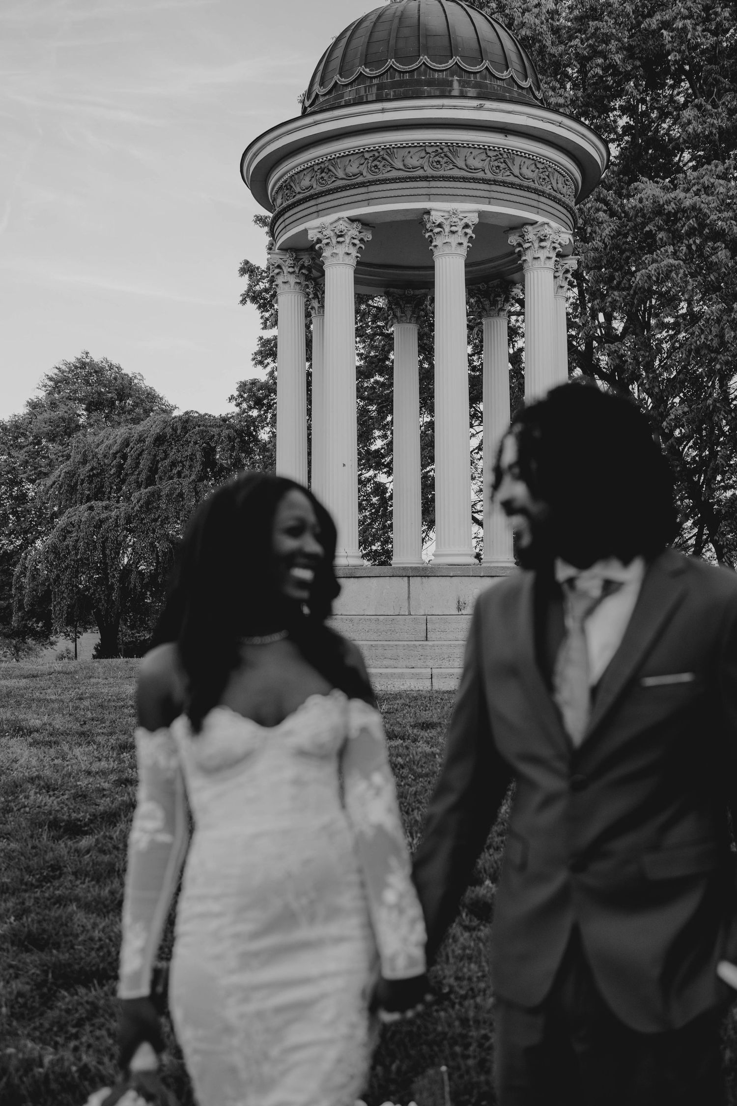Black and white photo of a smiling bride and groom holding hands in front of a classical rotunda gazebo.