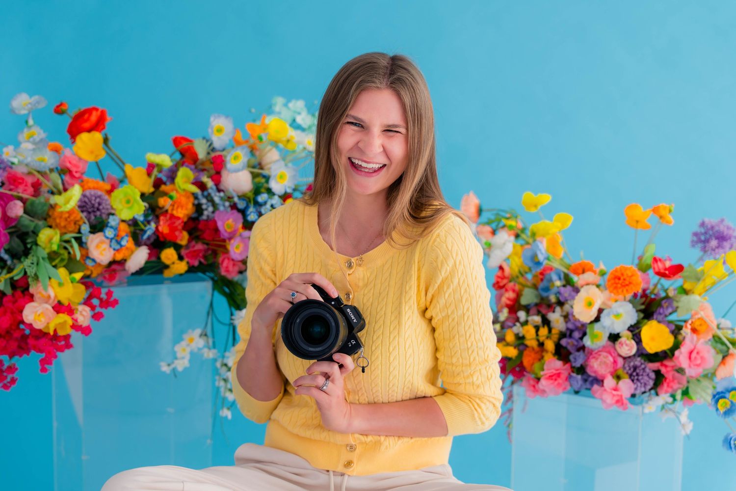 Laughing female photographer holding camera surrounded by colorful flowers on blue backdrop.
