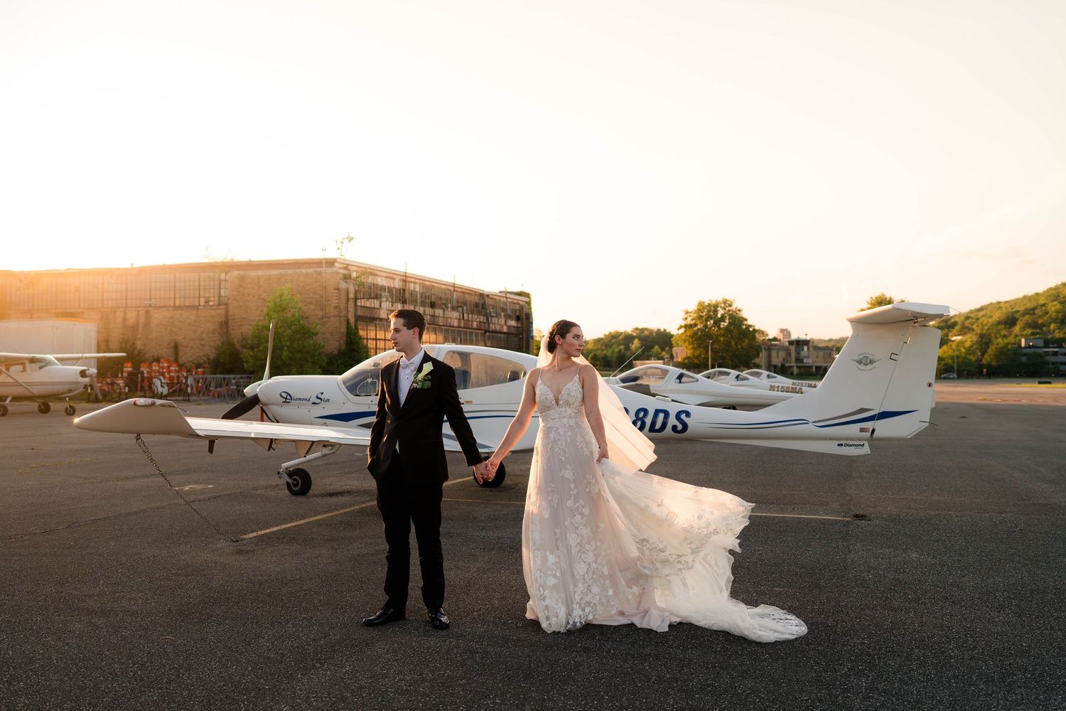 Bride and groom hold hands on airport tarmac at golden hour with small white plane behind them.