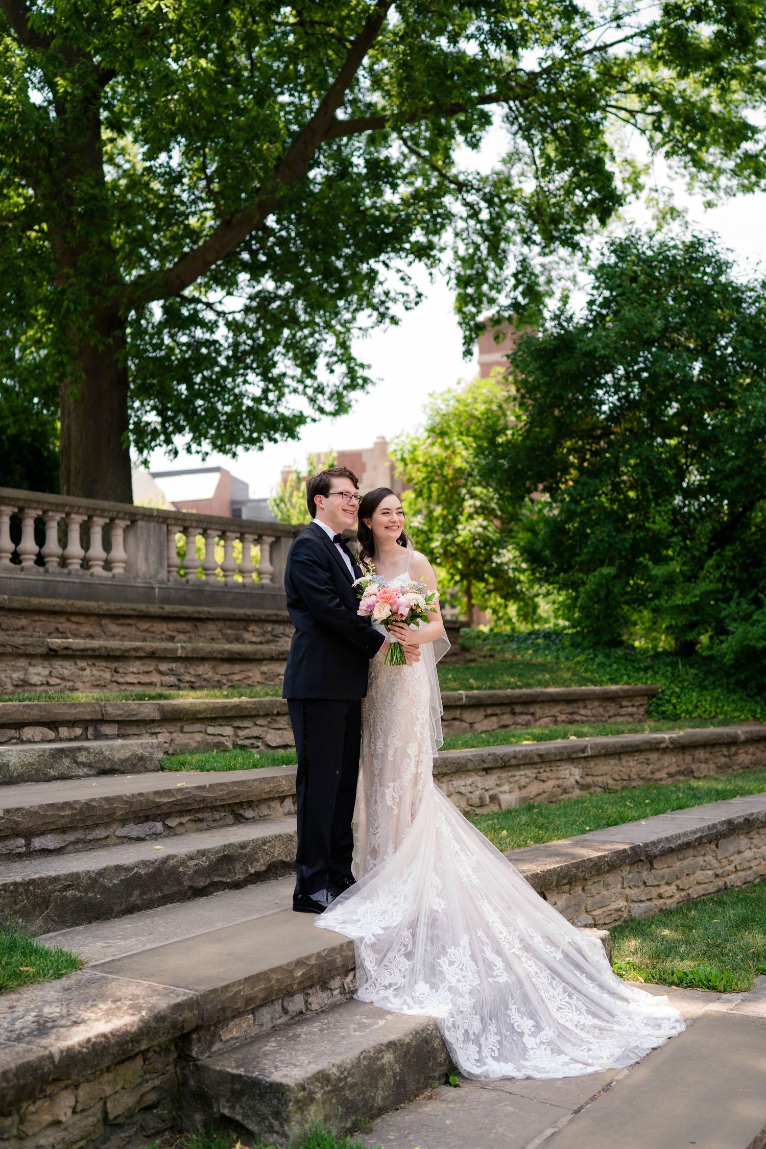 Bride and groom pose on stone steps beneath large tree at elegant outdoor venue.