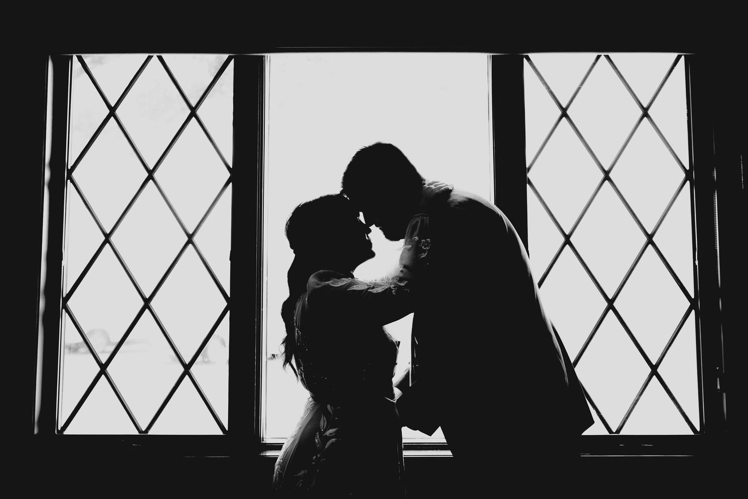 Romantic black and white silhouette of bride and groom nearly kissing before diamond-patterned window light.