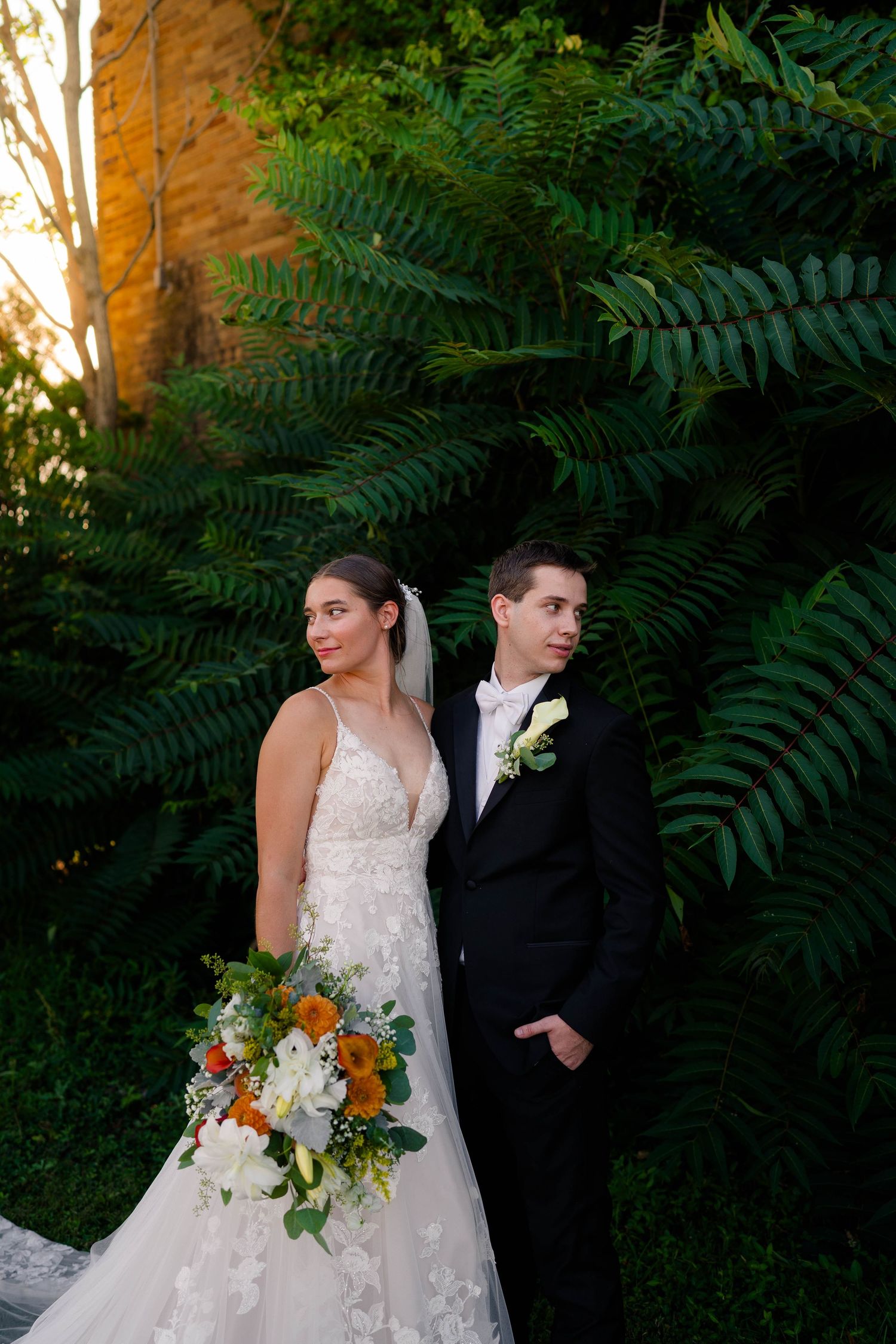 Bride in floral lace gown with colorful bouquet and groom in black tux beside tropical ferns.