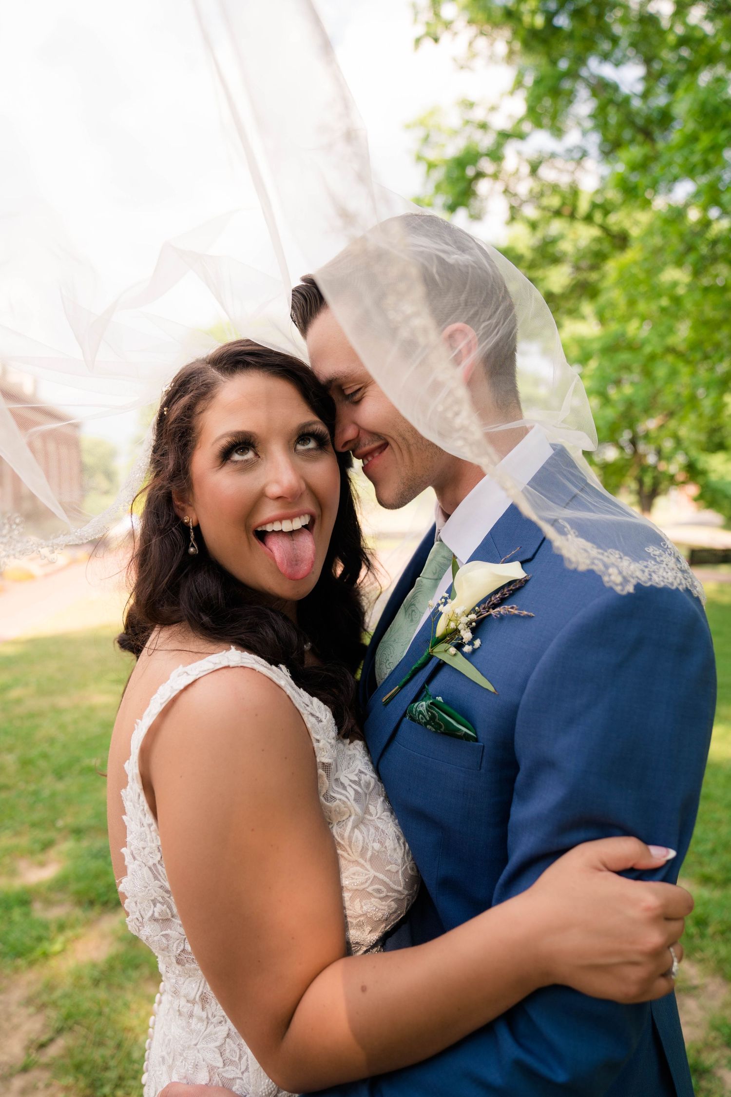 Joyful bride sticking out tongue embraces groom in blue suit under wedding veil outdoors.