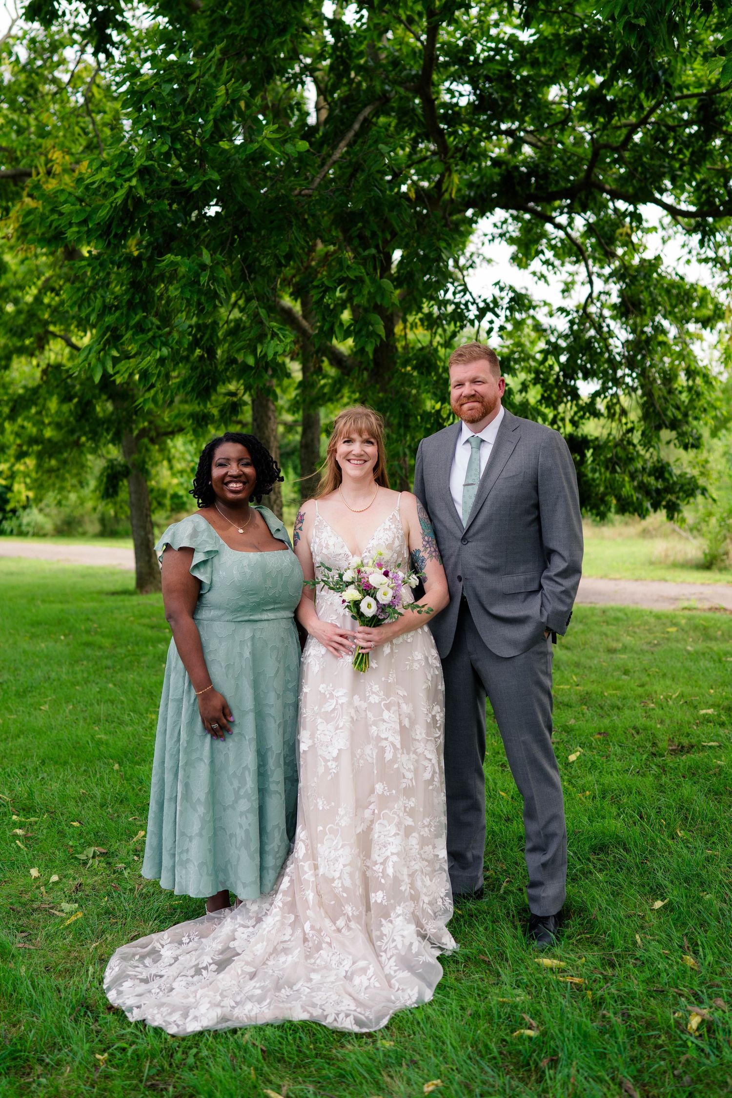 Bride in lace gown poses with bridesmaid in sage dress and groomsman in gray suit outdoors.
