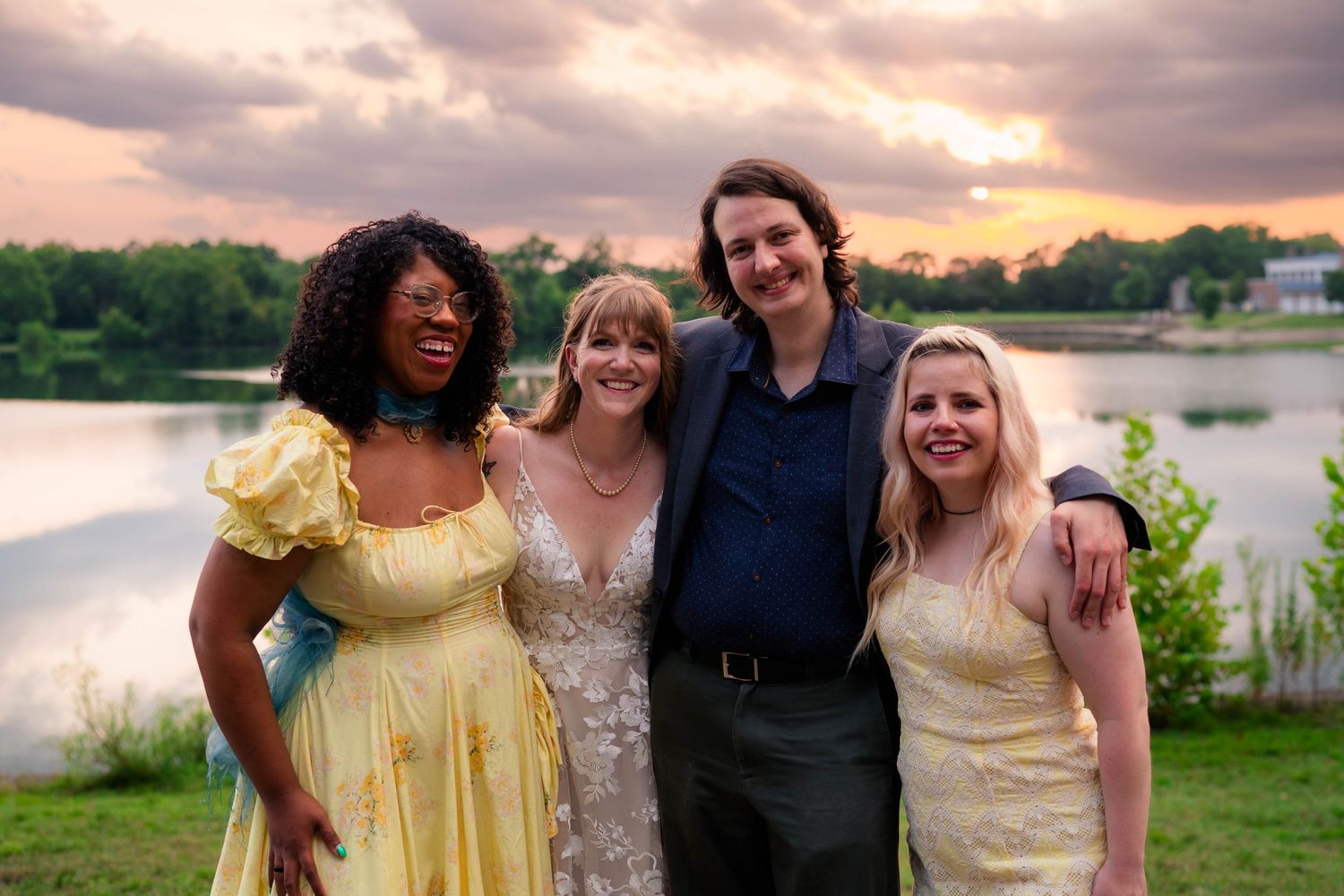 Four smiling wedding guests pose together in yellow dresses near a lake at sunset.